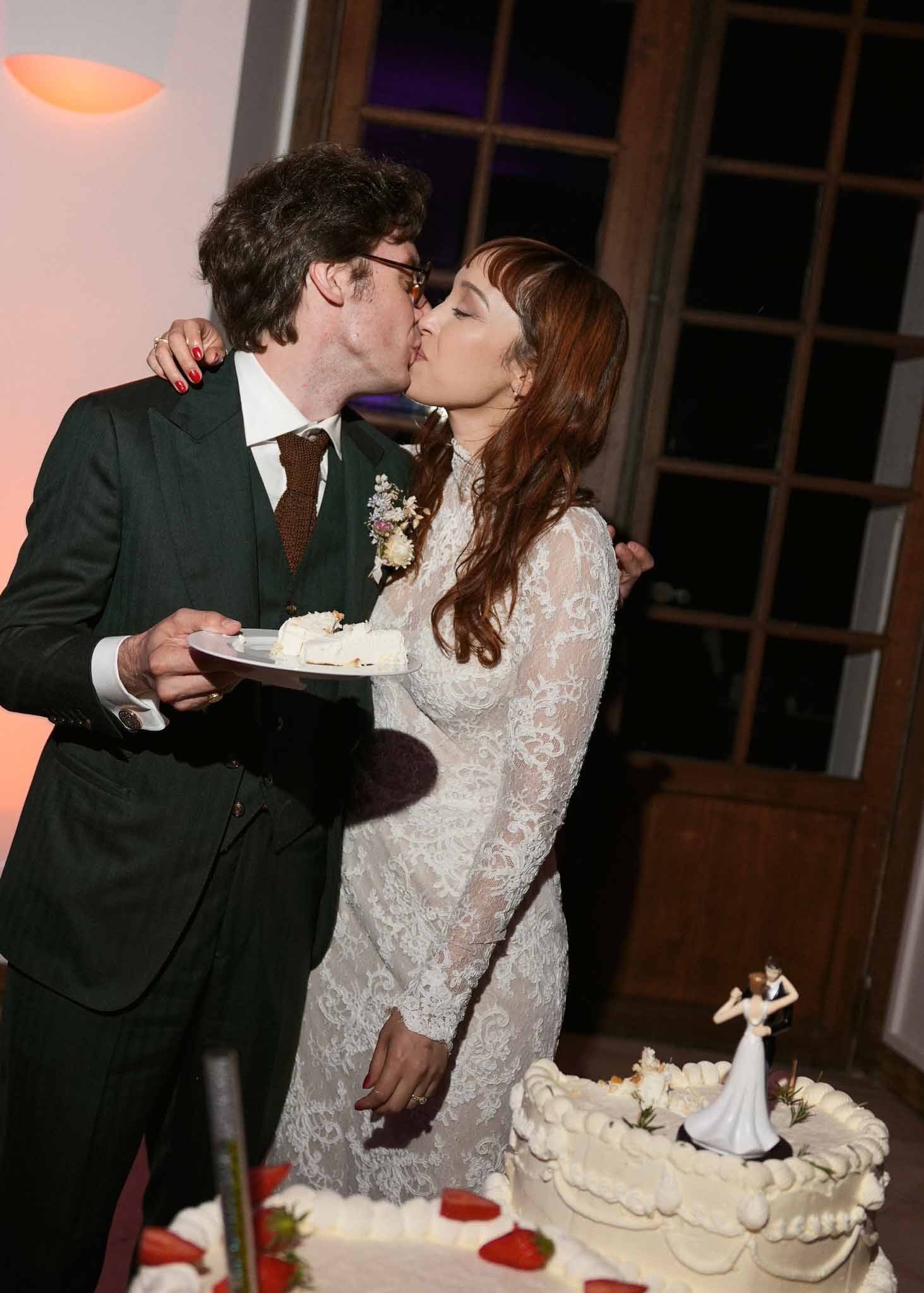 Bride and groom kissing during cake cutting at indoor wedding reception with tiered white cake