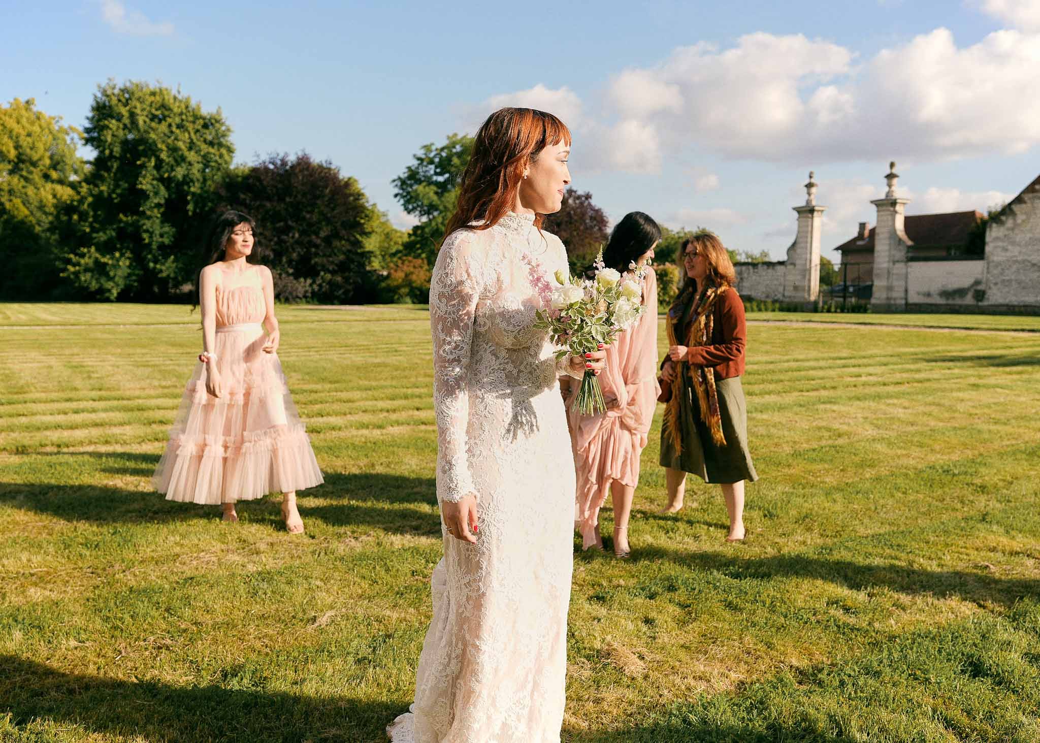 Bride in ivory lace gown with bouquet on country estate grounds with female guests in background