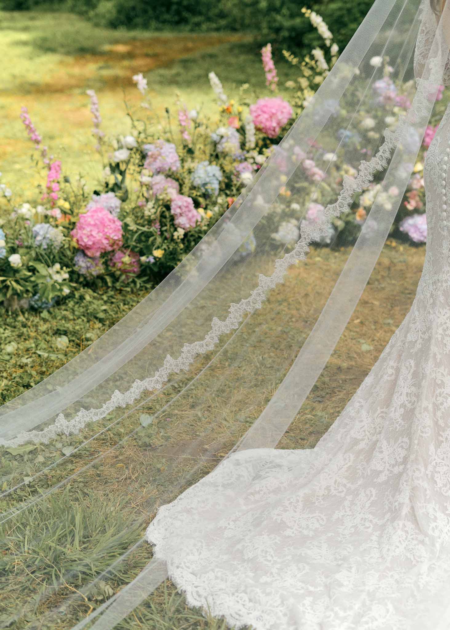 Bride's ivory lace-trimmed veil and train in cottage garden with pink peonies and lavender flowers