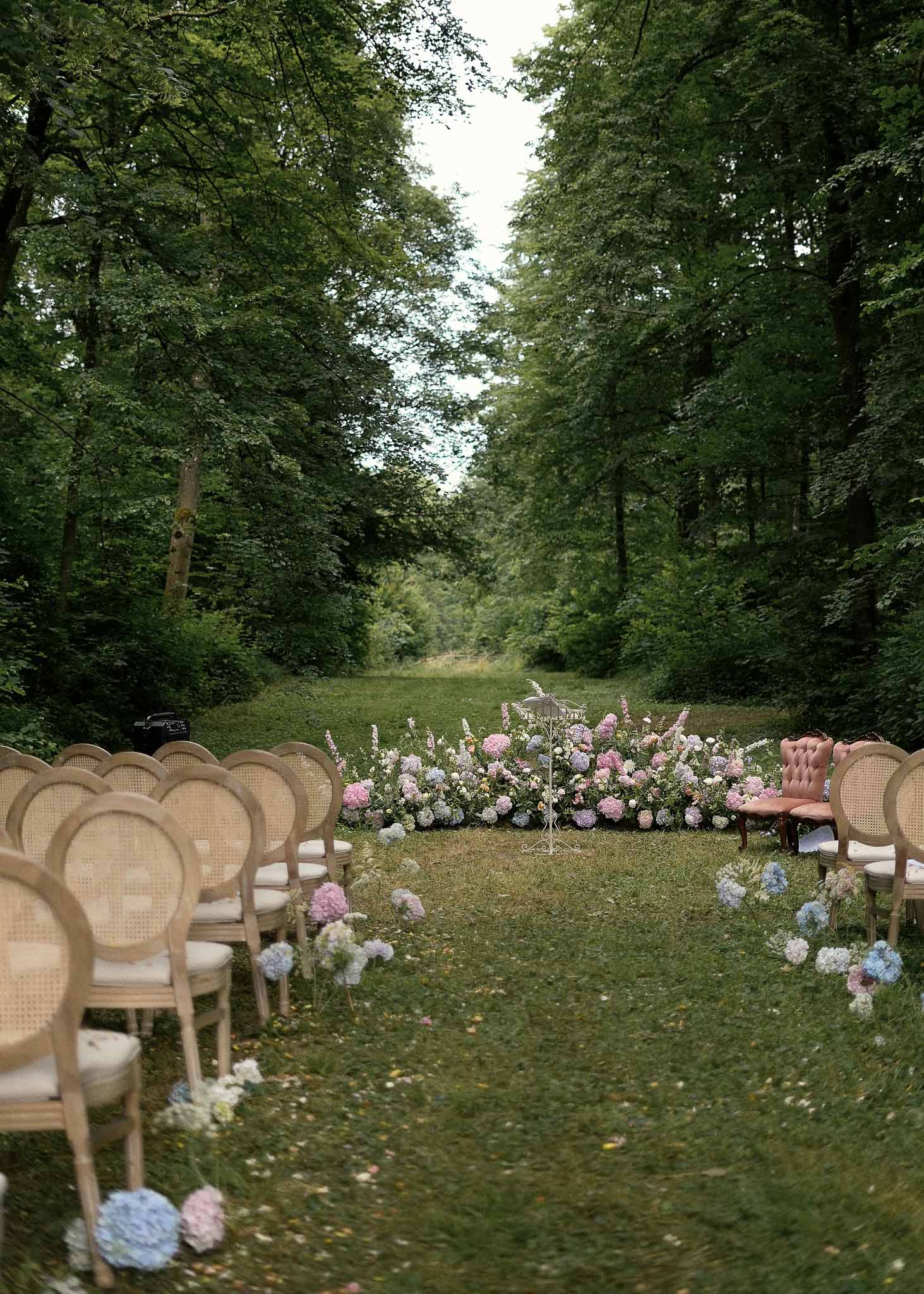 Outdoor wedding ceremony setup with wooden chairs and hydrangea aisle arrangements in woodland clearing