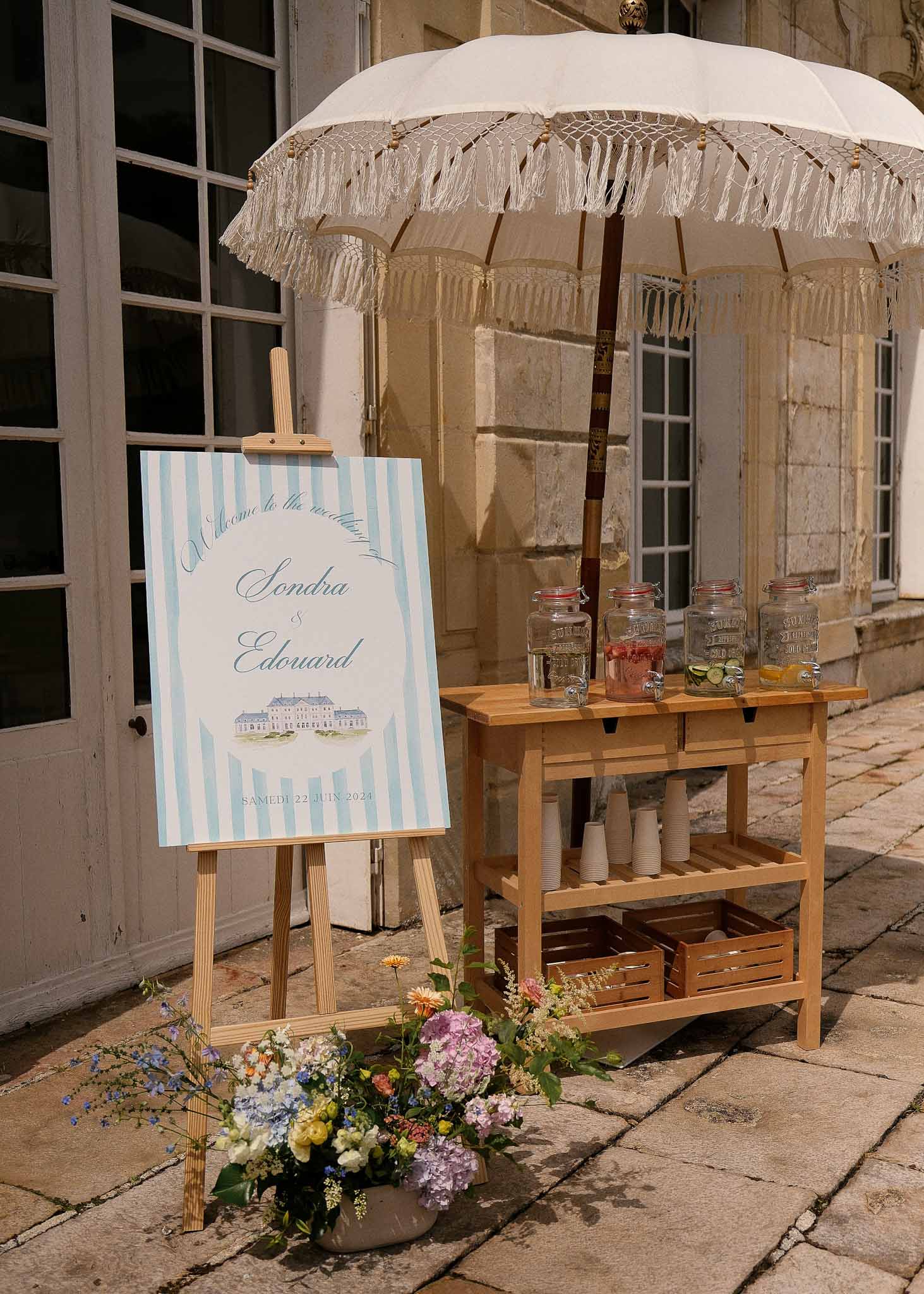 Welcome cocktail station with drinks dispensers and florals in classical French château courtyard