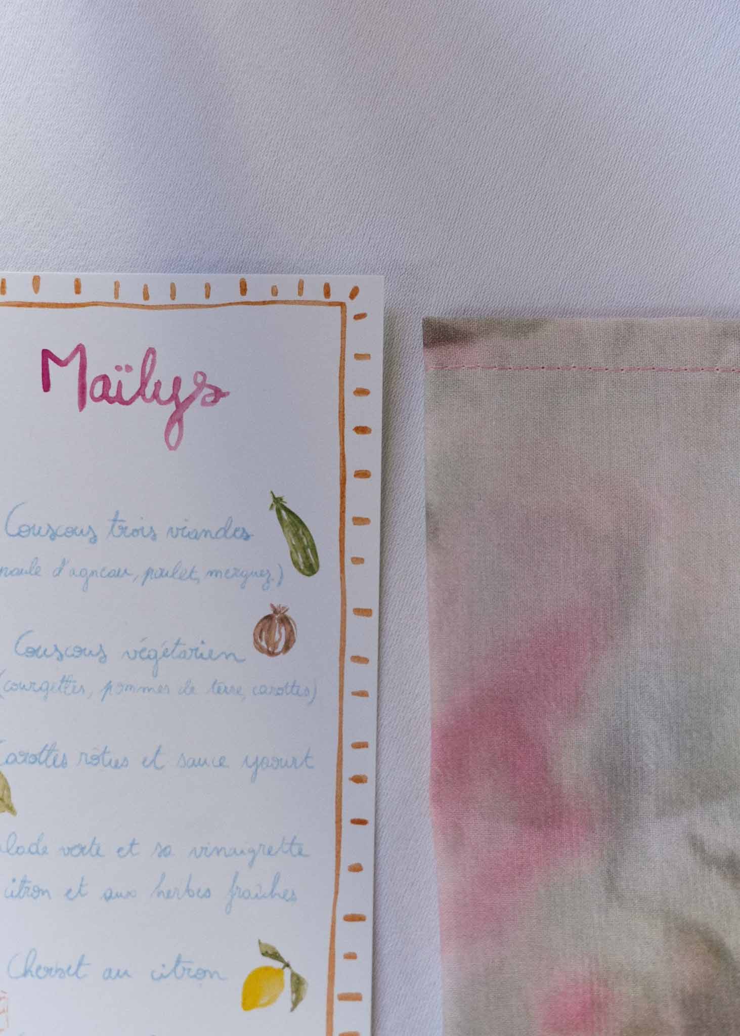 Illustrated watercolour menu card with guest name and vegetable drawings beside painted napkin on white linen