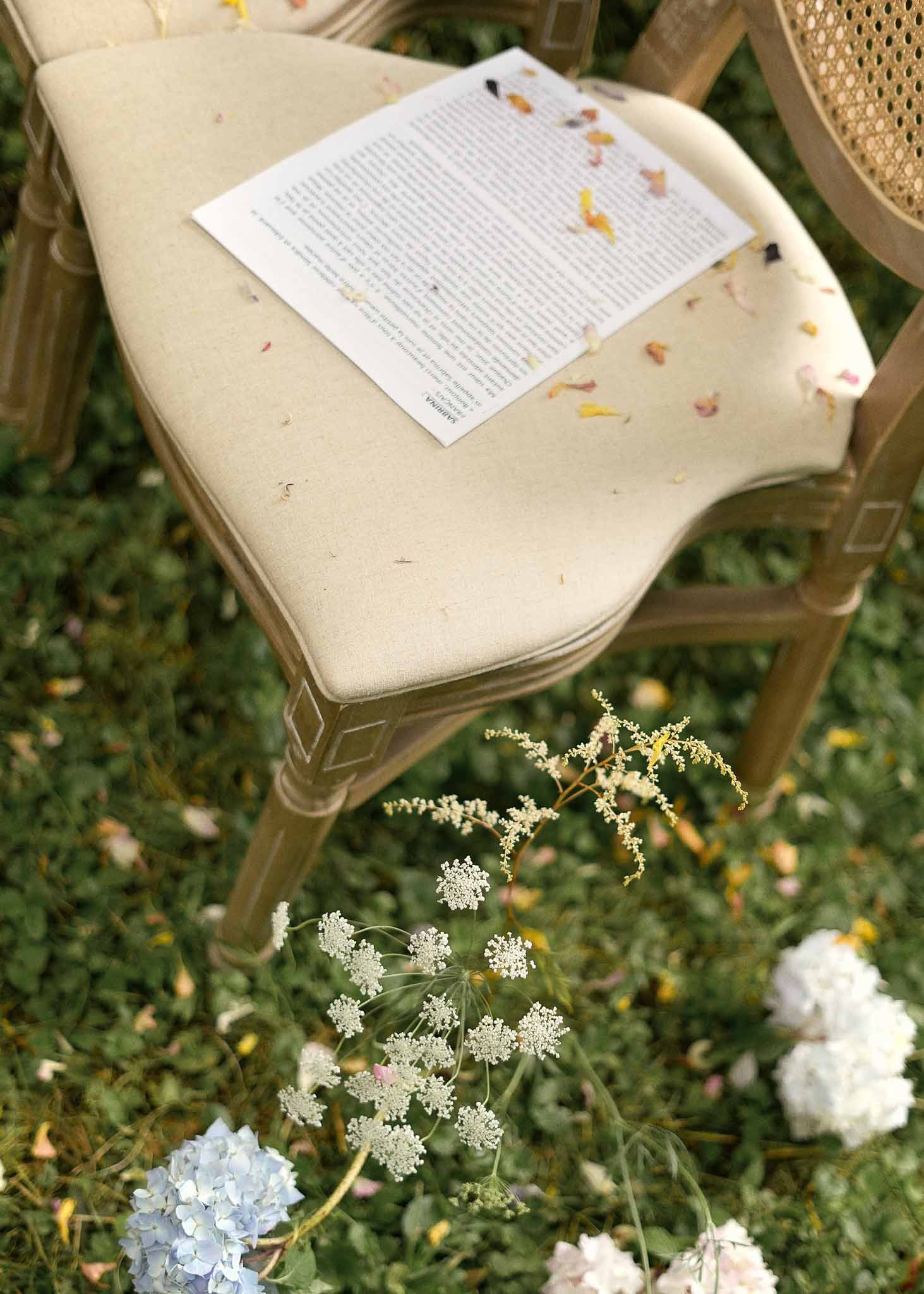 Wedding ceremony program with dried flower petals on wooden chair in garden setting