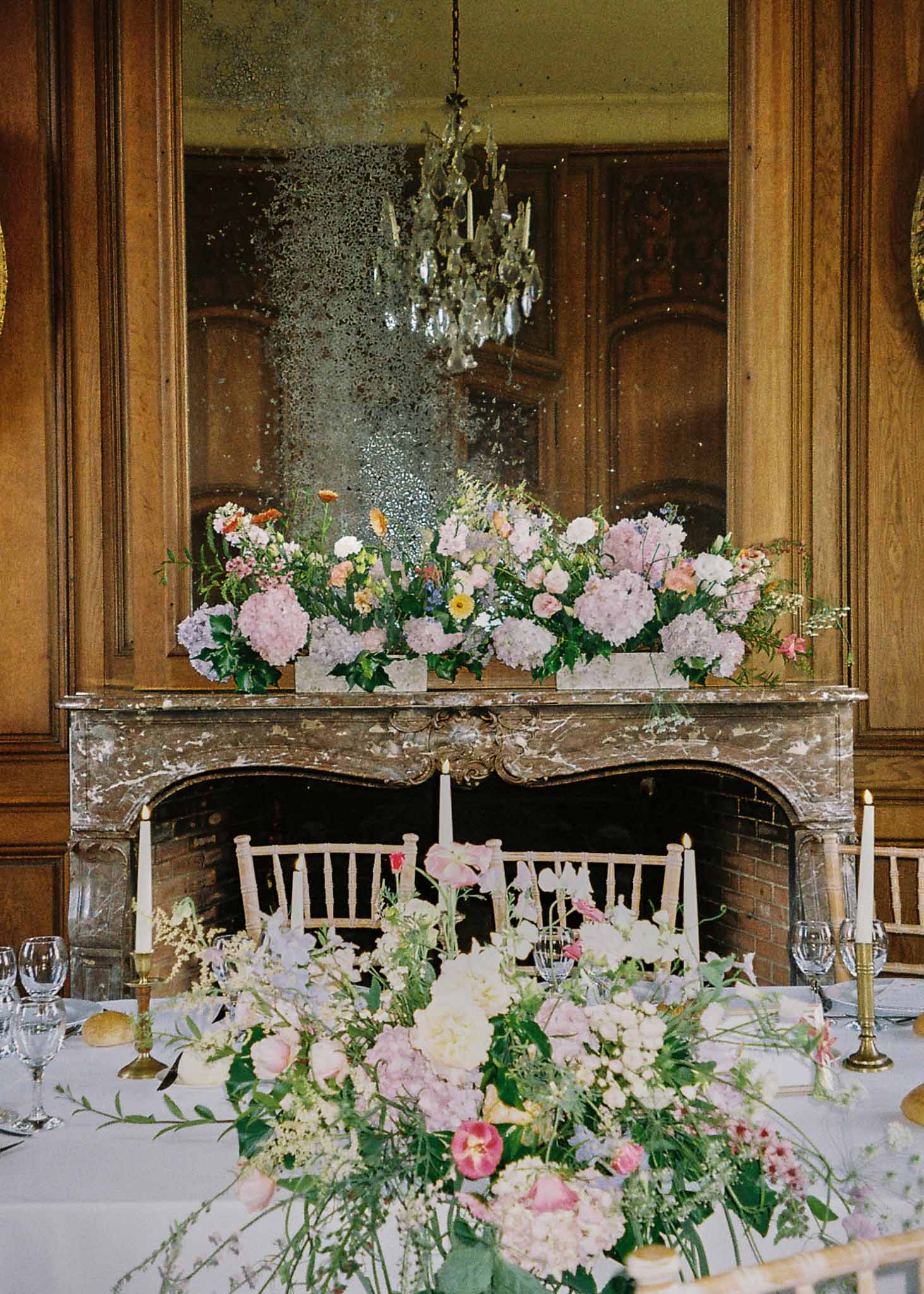 Reception dining setup with floral arrangements in ornate period room with marble fireplace