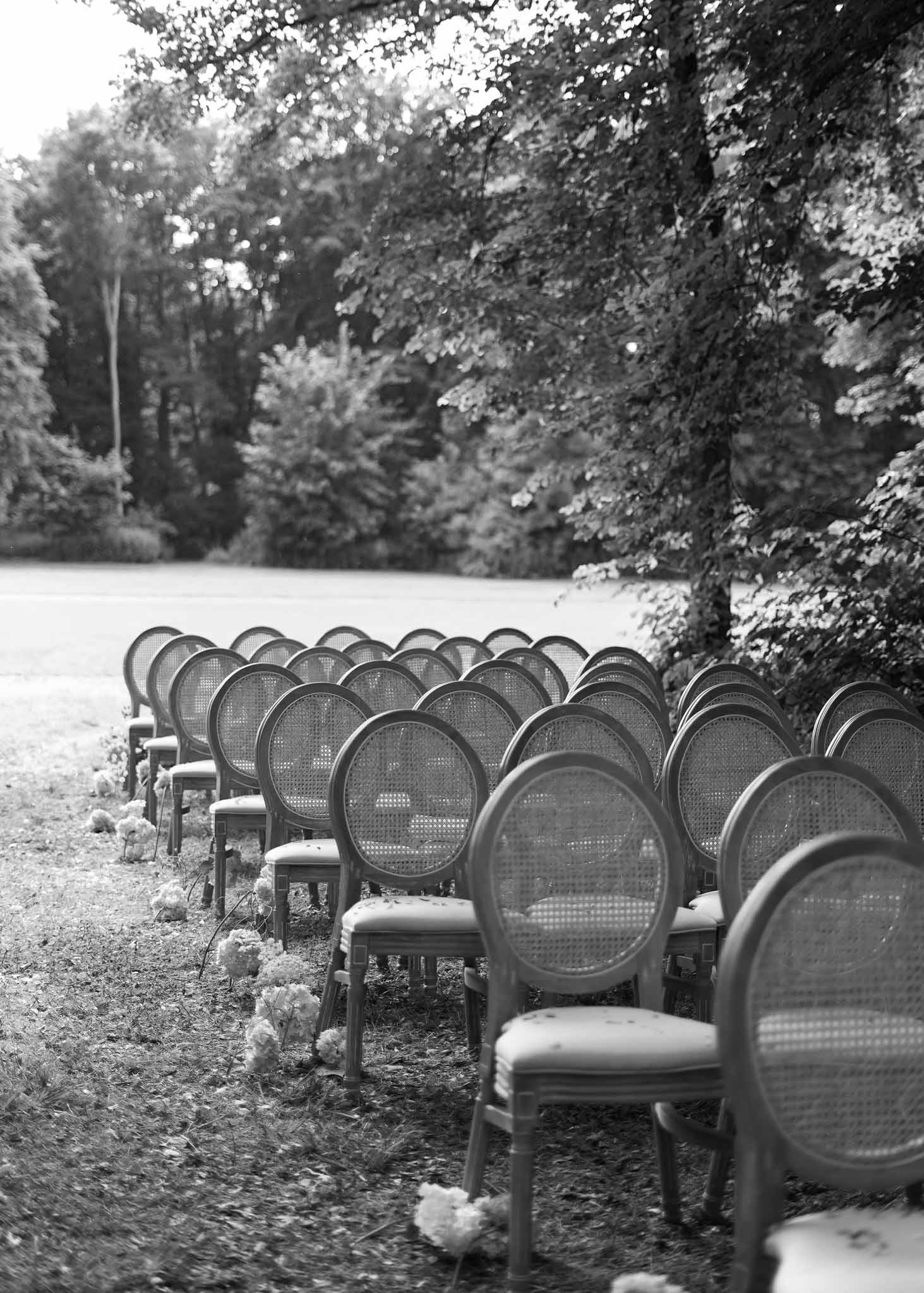 Outdoor wedding ceremony setup with Louis XVI chairs and floral aisle markers in wooded clearing