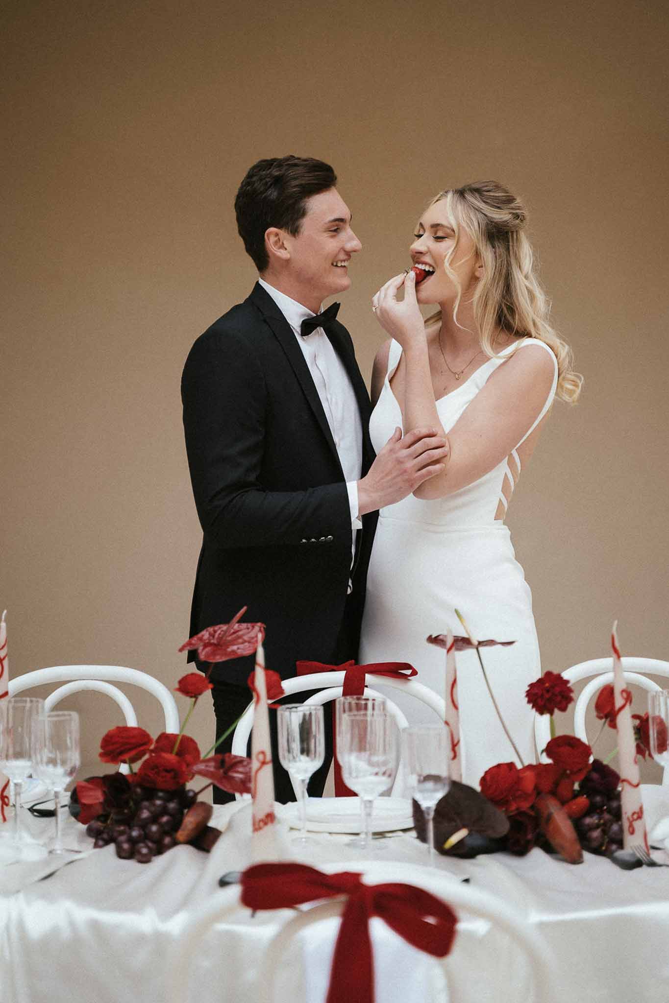 Groom feeding bride strawberry at sweetheart table with red anthurium and dark calla lily runner