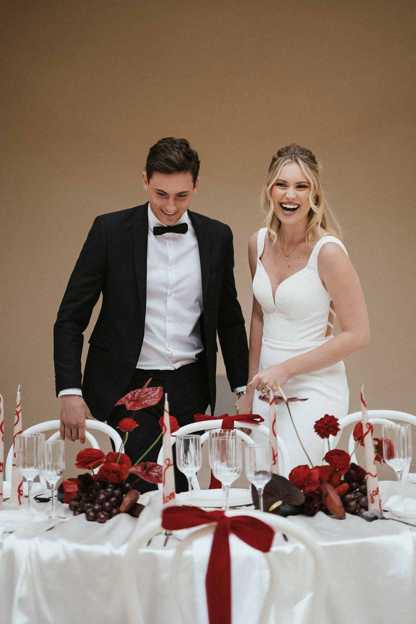 Bride and groom laughing behind reception table with deep red and burgundy floral arrangement, white taper candles, and re...