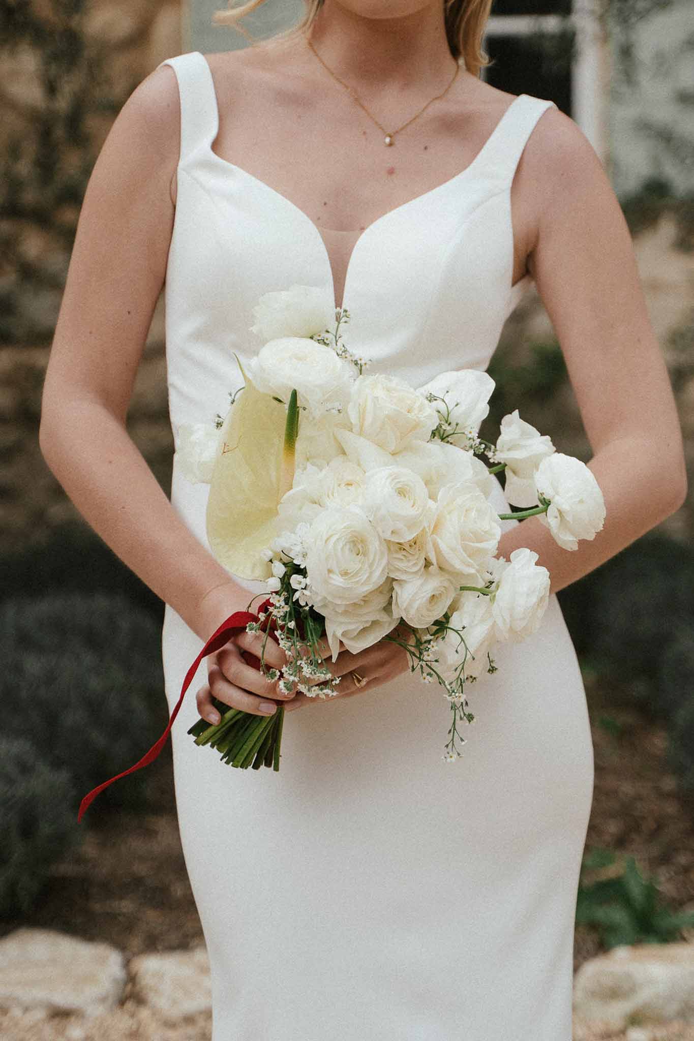 Close-up of bride holding ivory rose and ranunculus bouquet tied with deep red velvet ribbon against white fitted gown