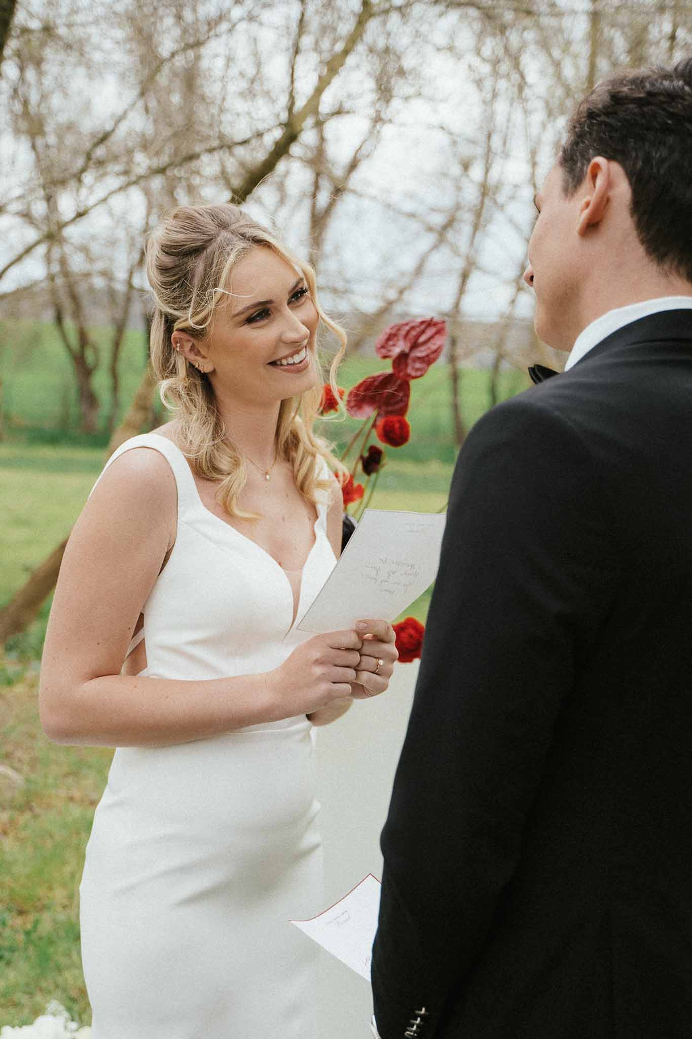 Bride in ivory V-neck gown reading handwritten vows to groom during outdoor ceremony with deep red anthurium bouquet