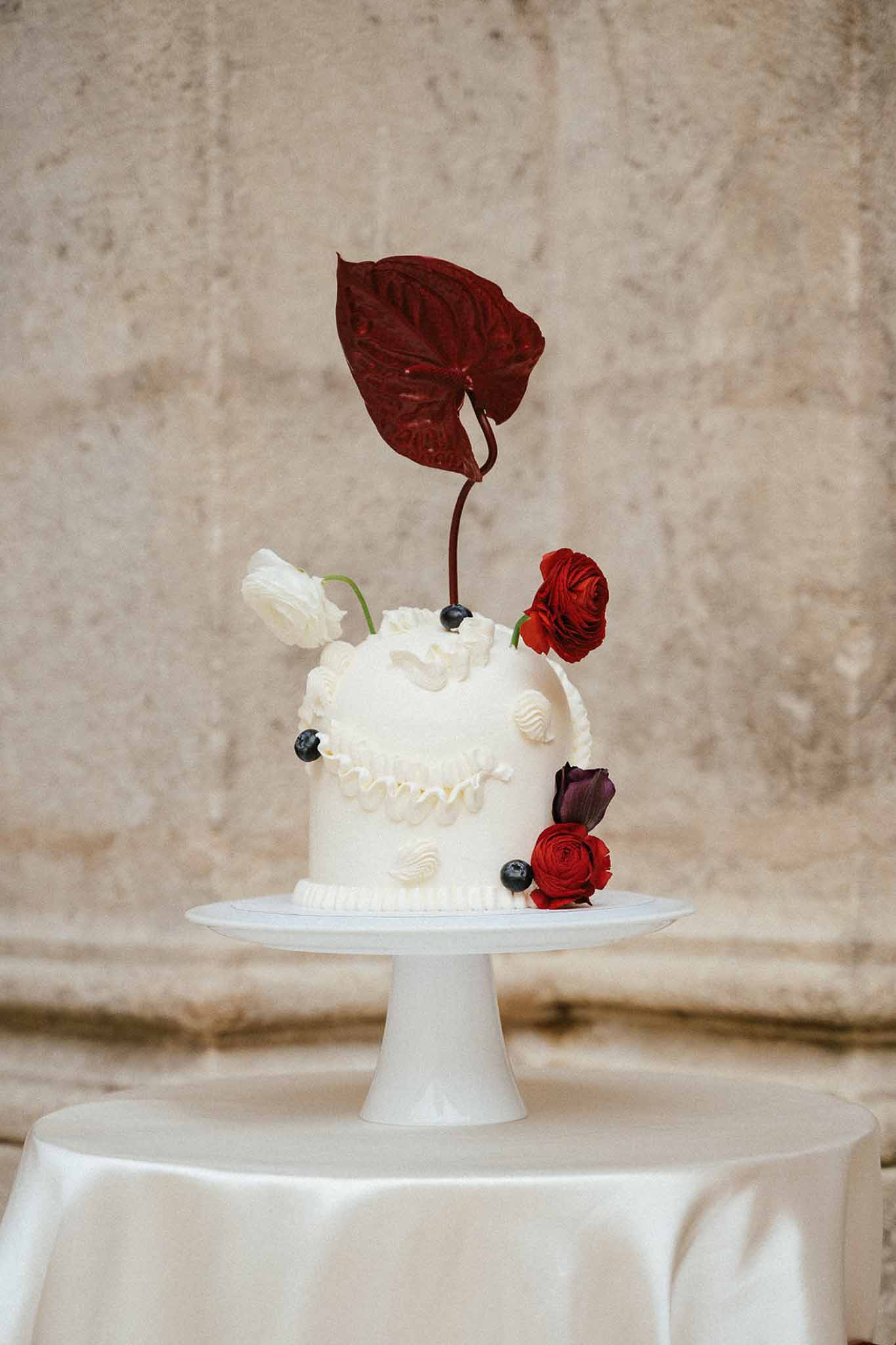 Single-tier white wedding cake with deep red ranunculus and burgundy anthurium on a pedestal stand