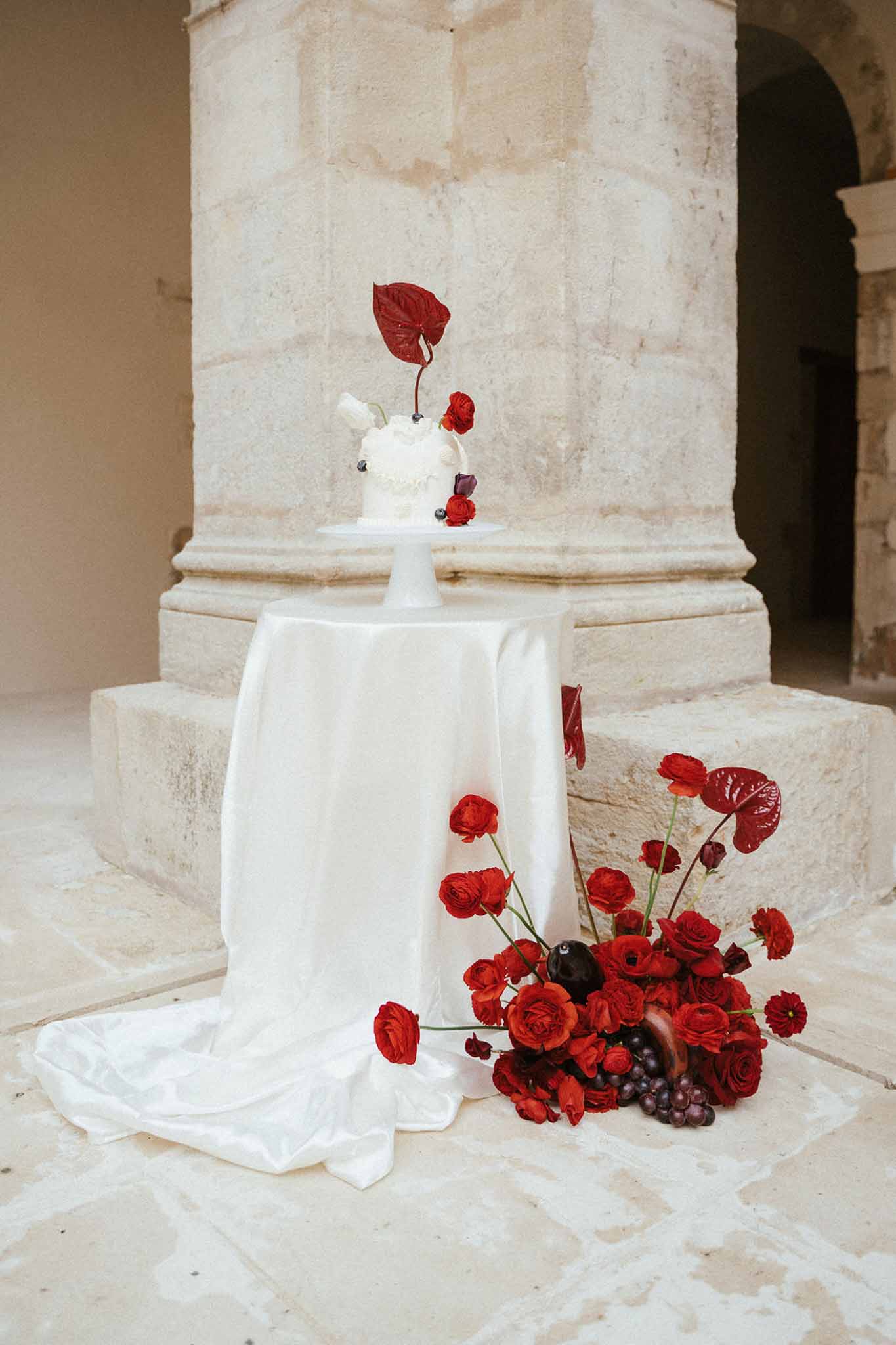 Red floral arrangement and white cake display on pedestal table in historic stone archway setting
