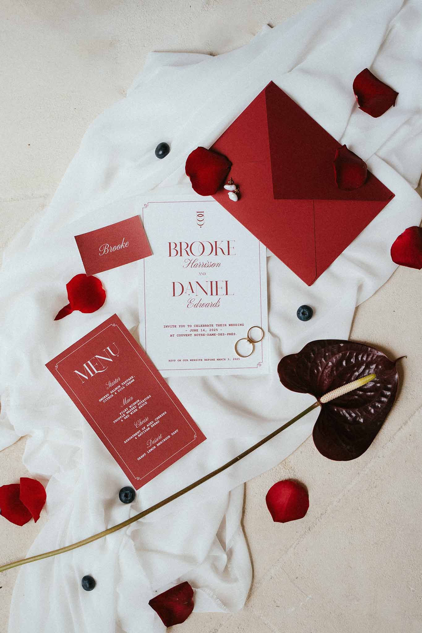 Stationery flat lay with deep red typography envelope and menu gold wedding bands red rose petals and burgundy anthurium