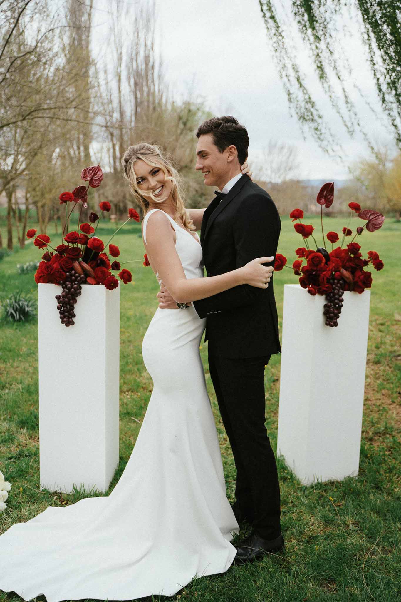 Couple embracing between white pedestals with deep red rose and anthurium arrangements