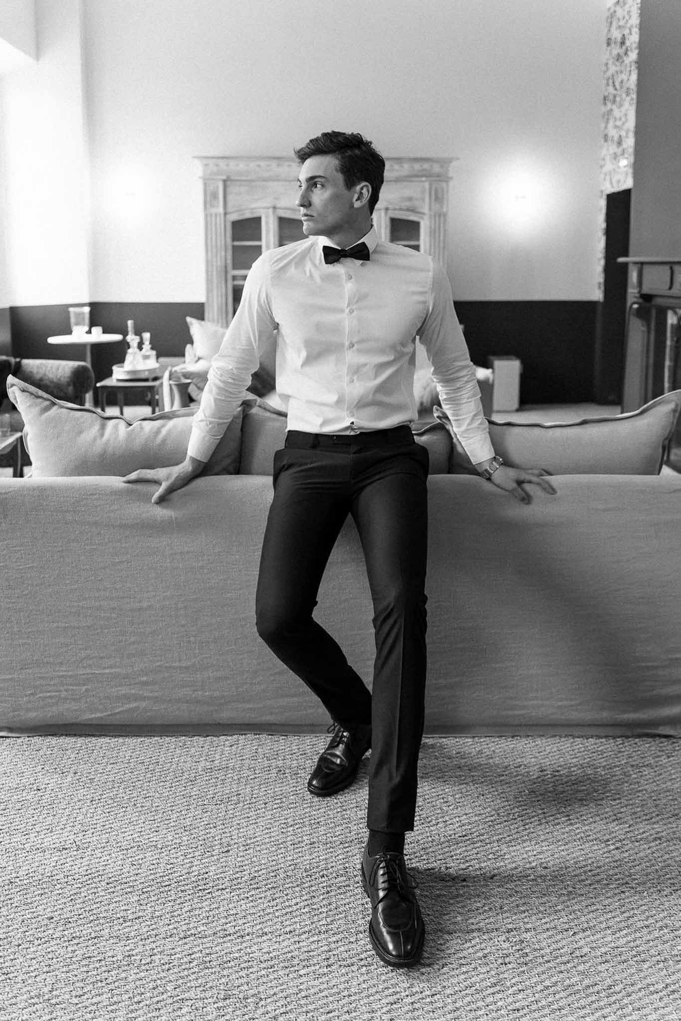 Black and white full-length portrait of groom leaning against sofa in formal chateau sitting room