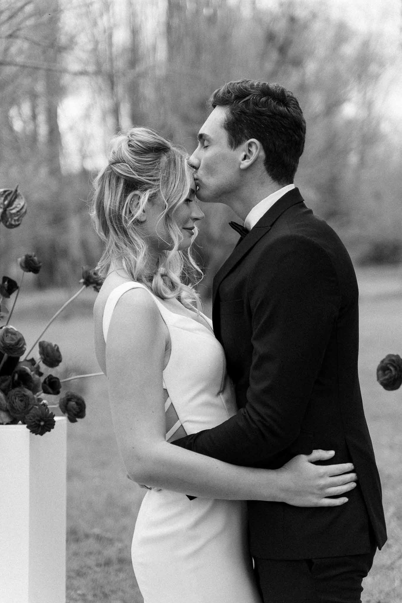Groom kisses bride on forehead beside modern floral arrangement with anthurium leaves in high-contrast B&W