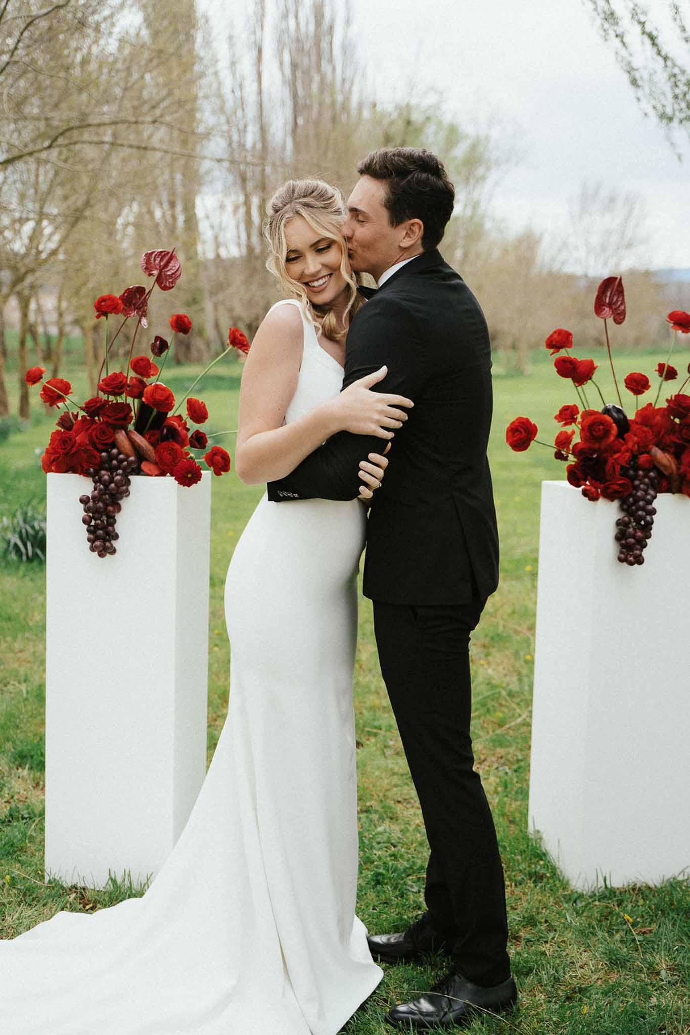 Groom kissing bride on cheek beside tall white pedestals topped with deep red rose and anthurium arrangements