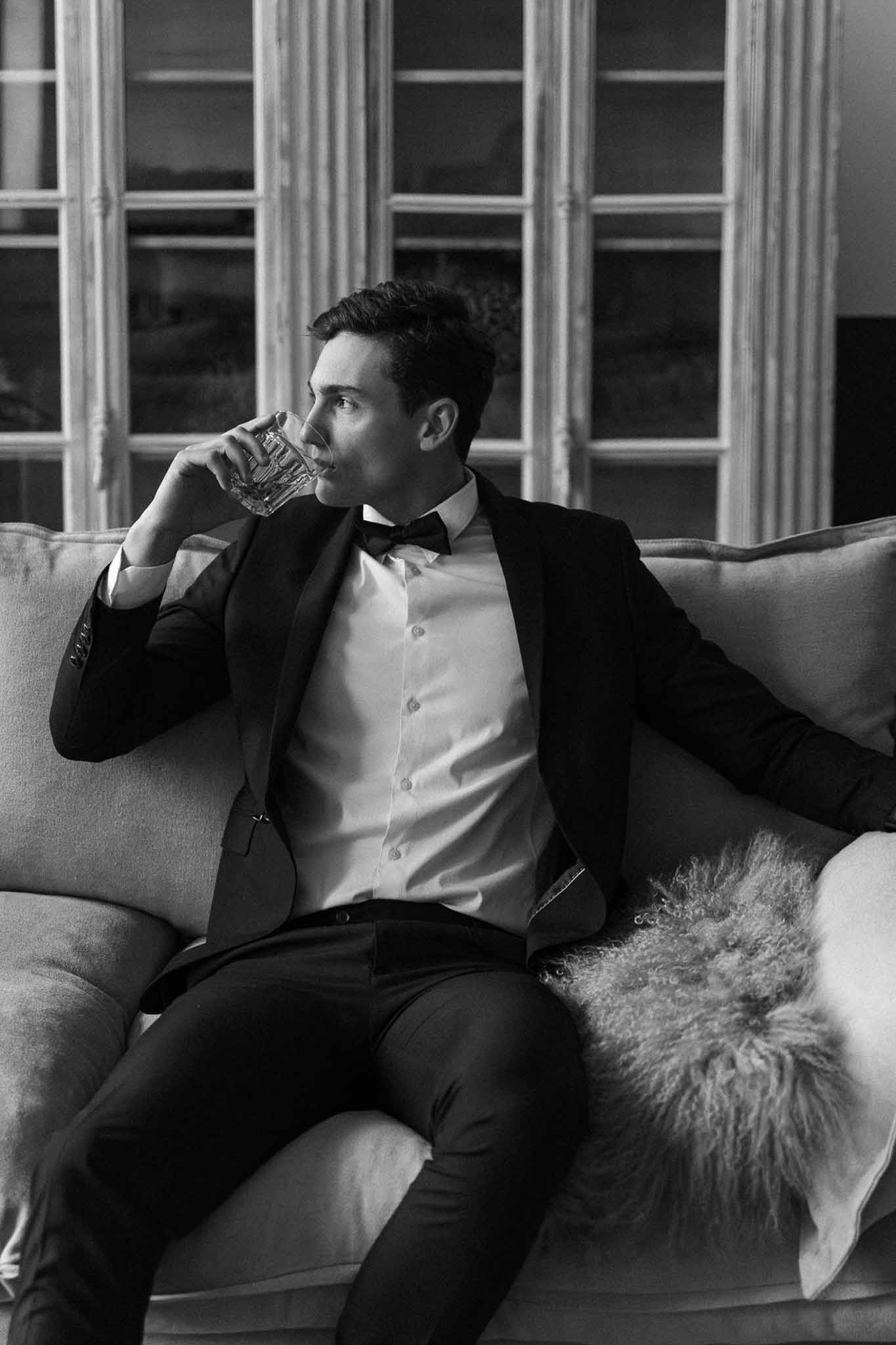 Black-and-white portrait of groom in tuxedo and bow tie seated on sofa drinking from crystal glass