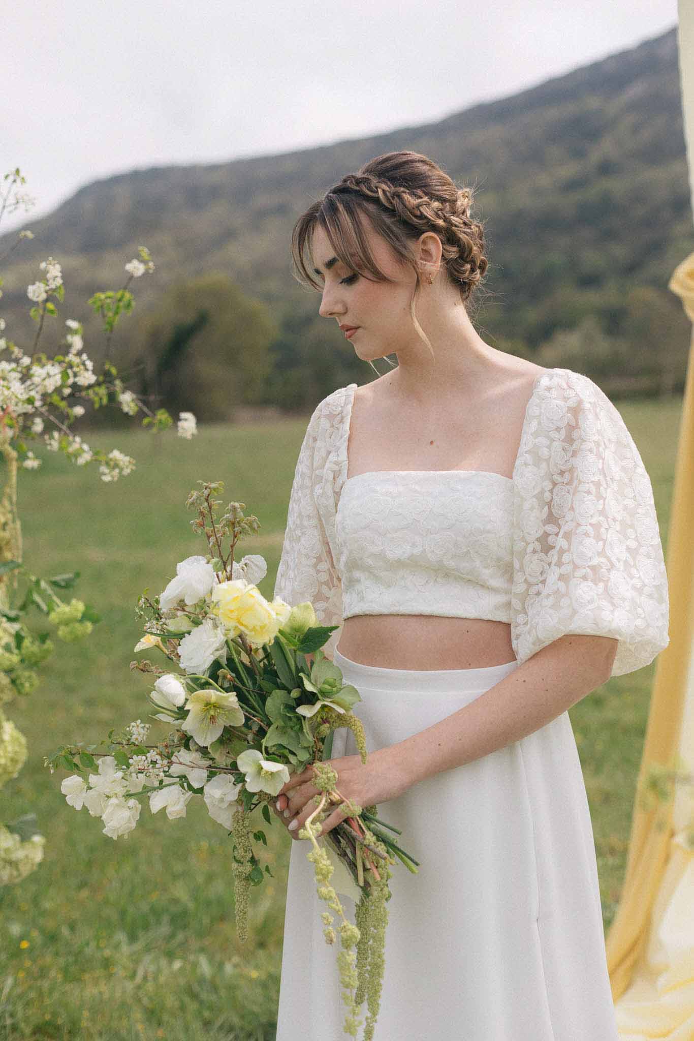 Bride in ivory lace crop top and white skirt holding bouquet in countryside meadow setting