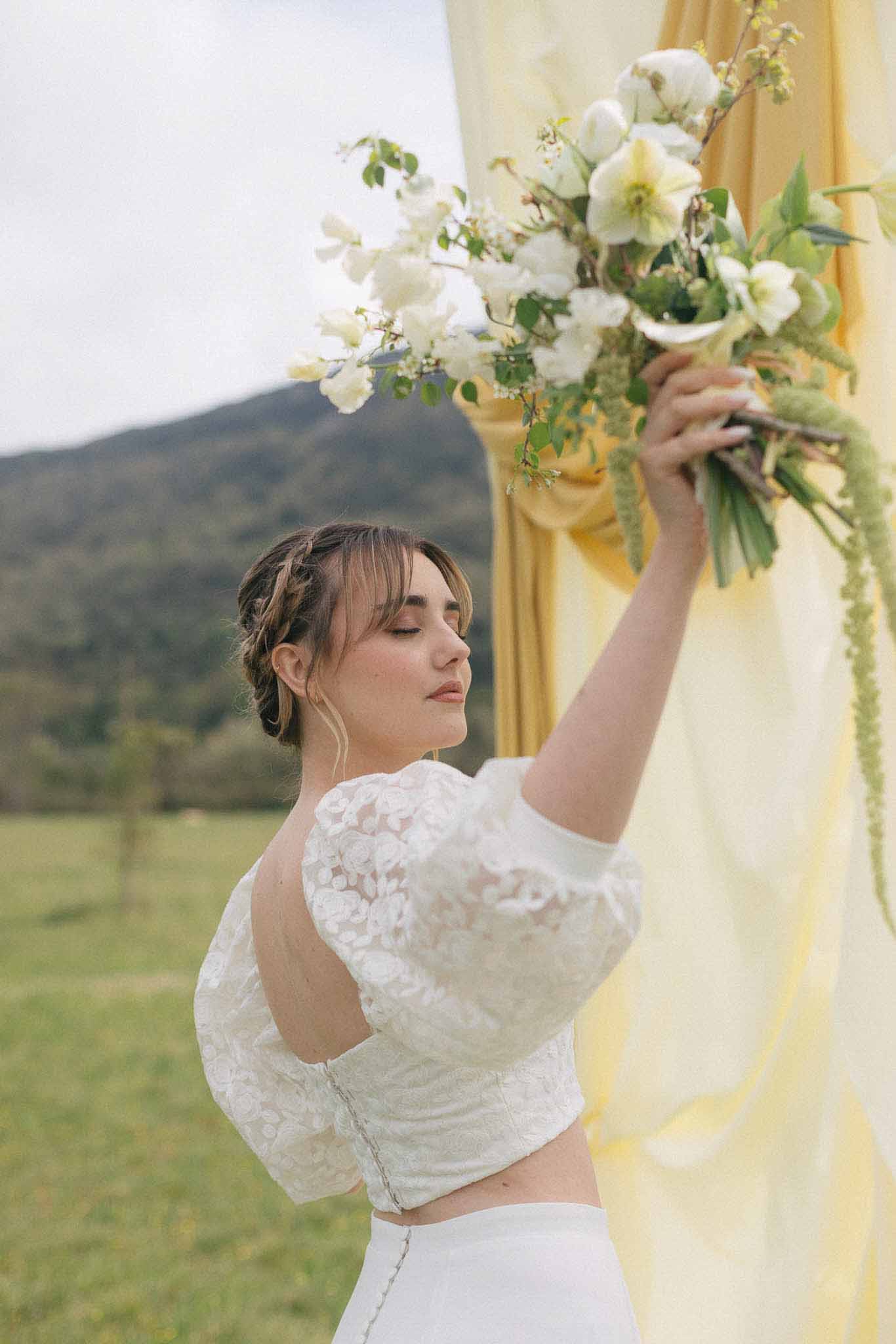 Bride in white lace crop top wedding dress holding white bouquet at outdoor mountain venue