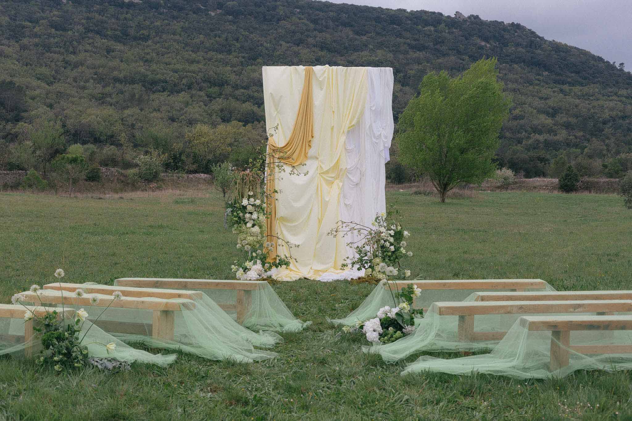 Outdoor wedding ceremony setup with draped arch and wooden benches in open field with hills