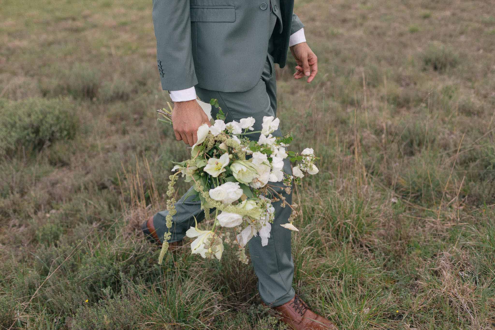 Groom in sage green suit holding white bridal bouquet with ranunculus and garden roses in outdoor field setting