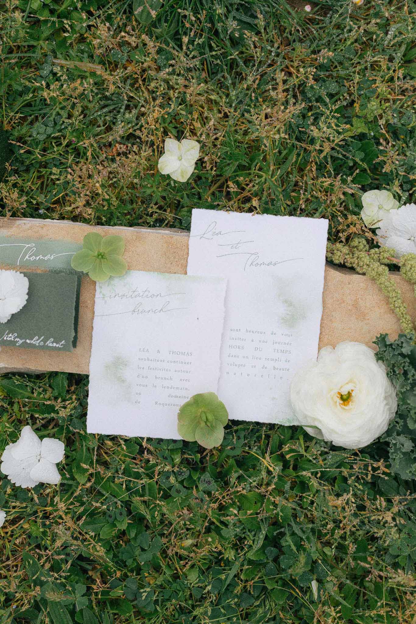 Wedding invitation flat lay with white flowers and sage green cards on grass