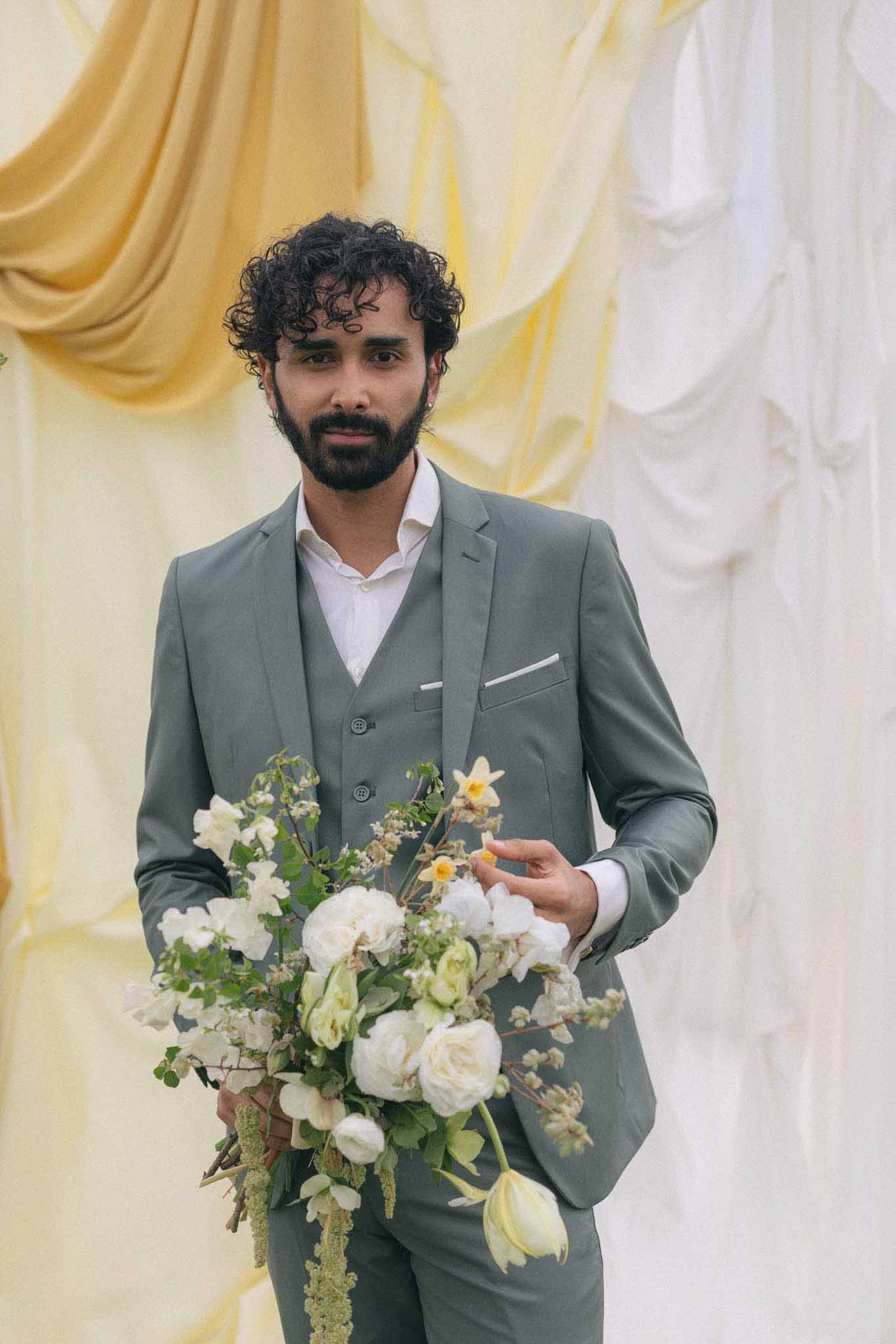 Groom in sage green suit holding bridal bouquet against draped fabric backdrop at wedding venue