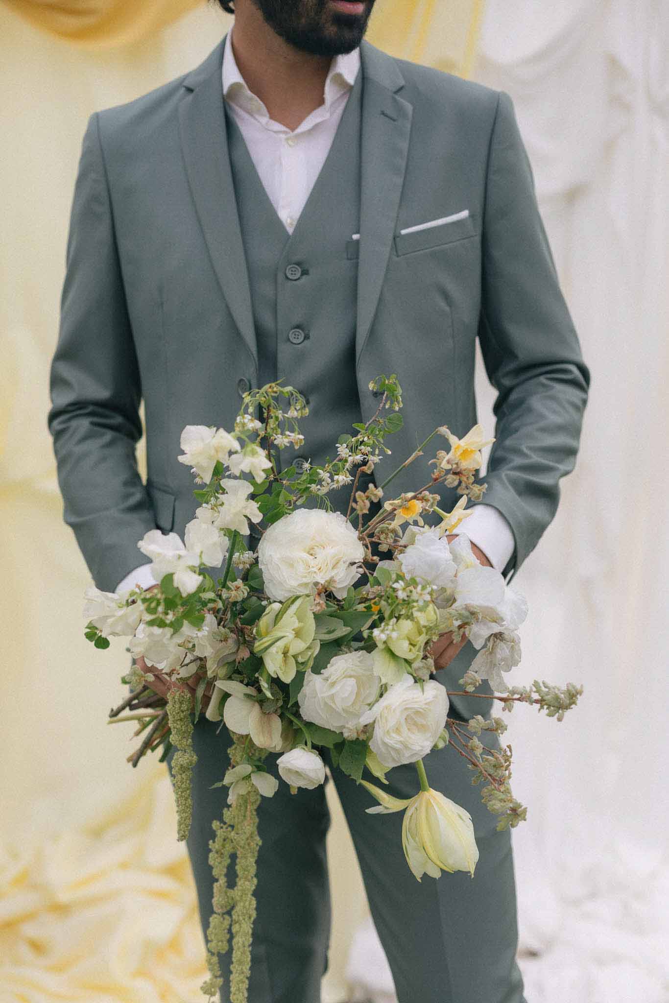 Groom in sage green suit holding cream and white bridal bouquet indoors