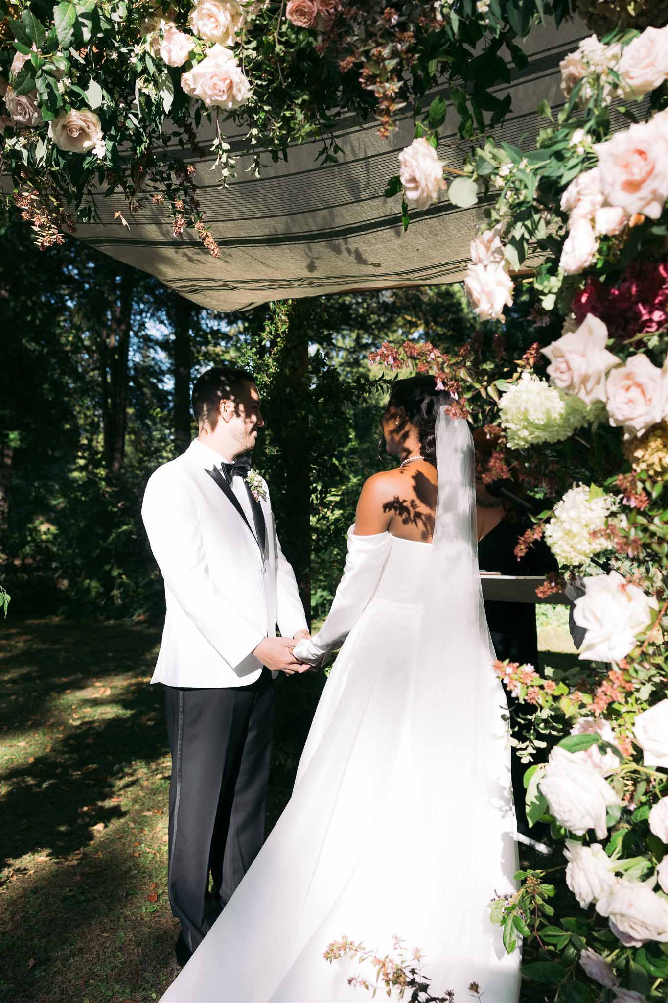 Bride and groom holding hands during outdoor ceremony beneath flower-decorated pergola in garden setting
