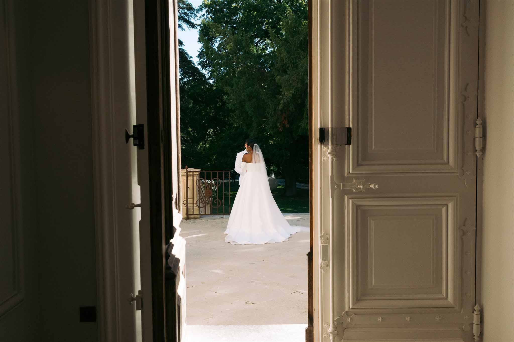 Bride in white dress and veil standing on stone terrace framed by French doors overlooking wooded landscape