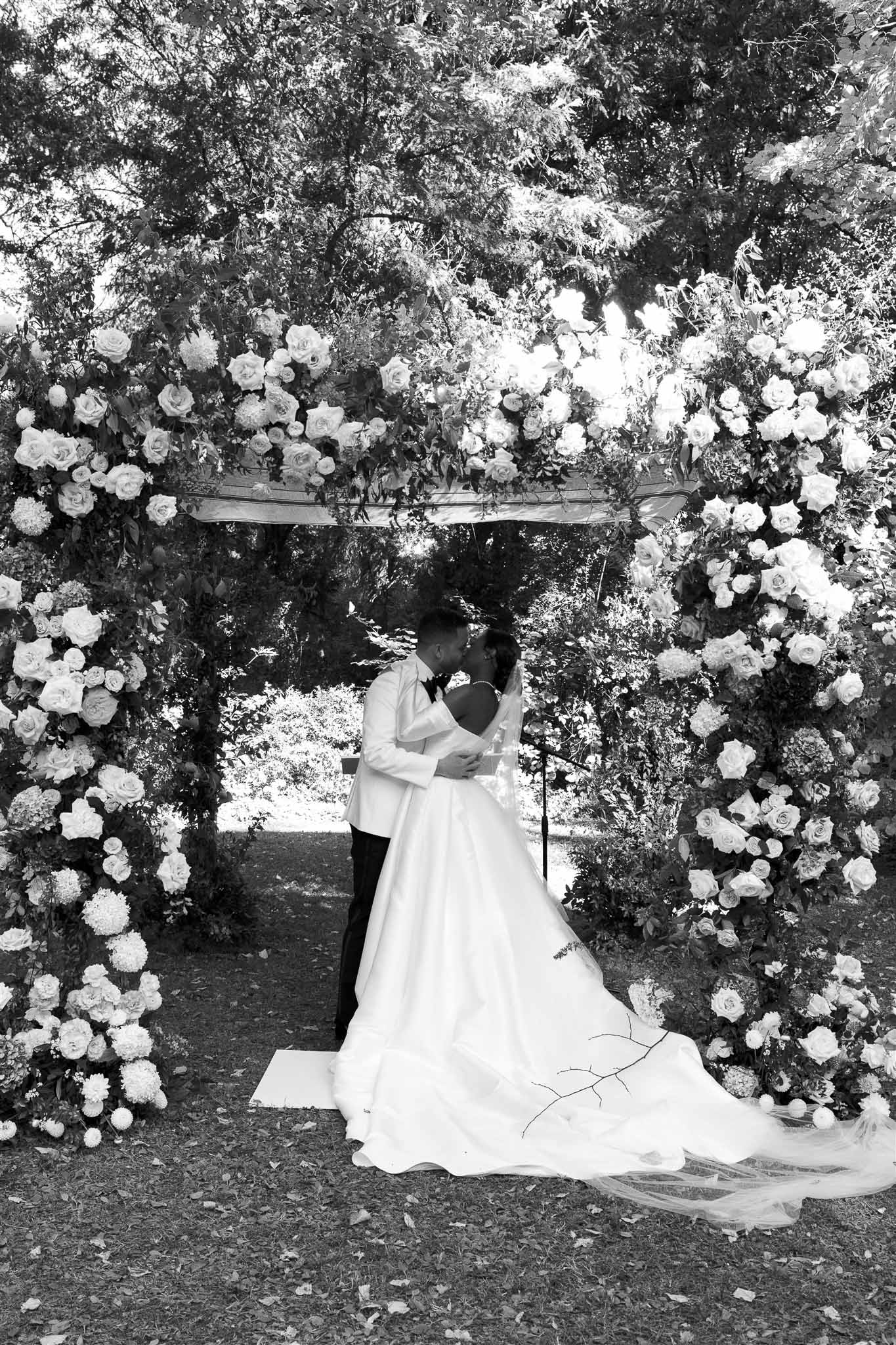 Bride and groom exchanging vows under rose-covered archway during outdoor garden wedding ceremony