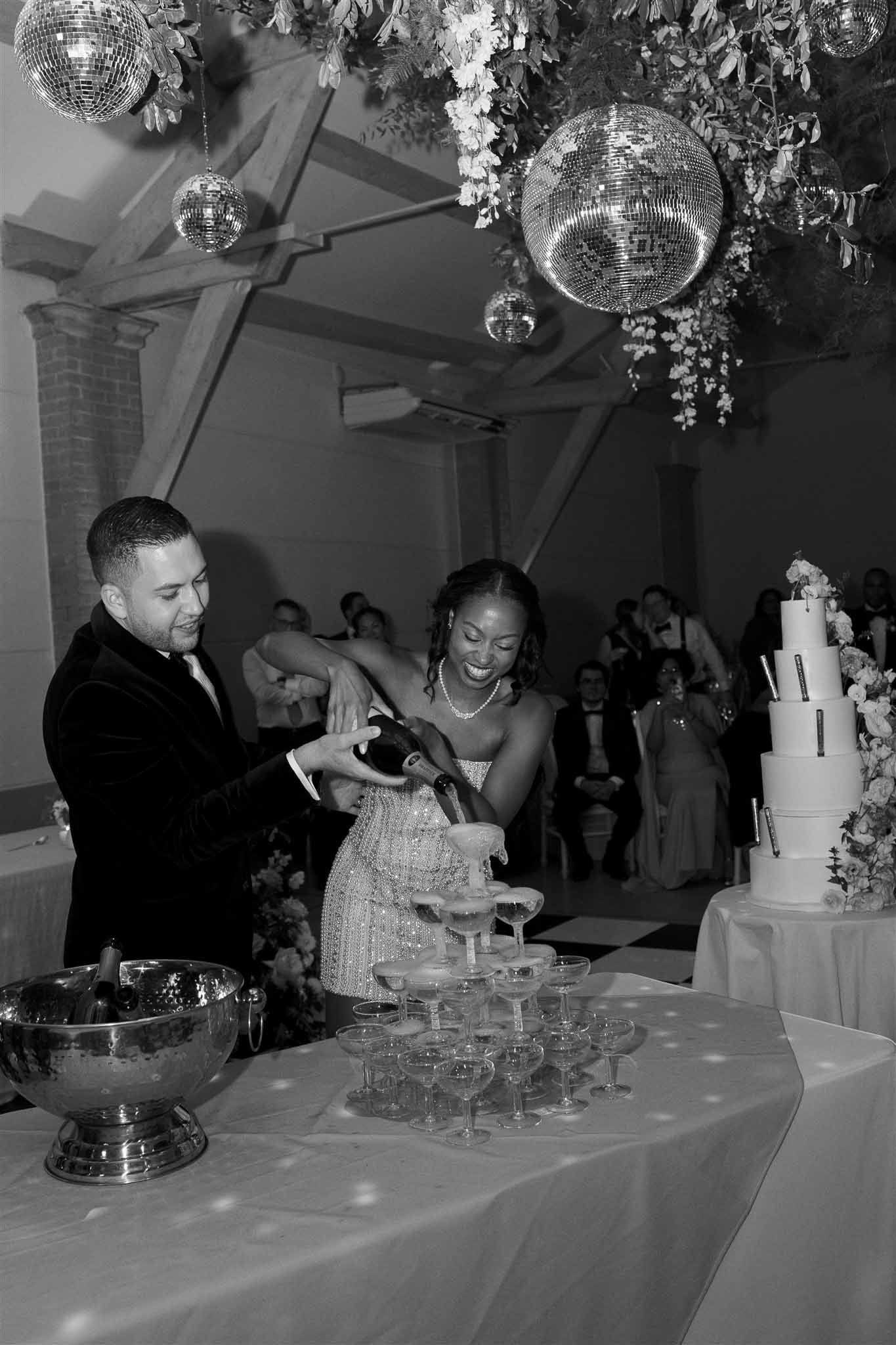 Couple pouring champagne tower at industrial modern wedding reception with disco balls and floral installations