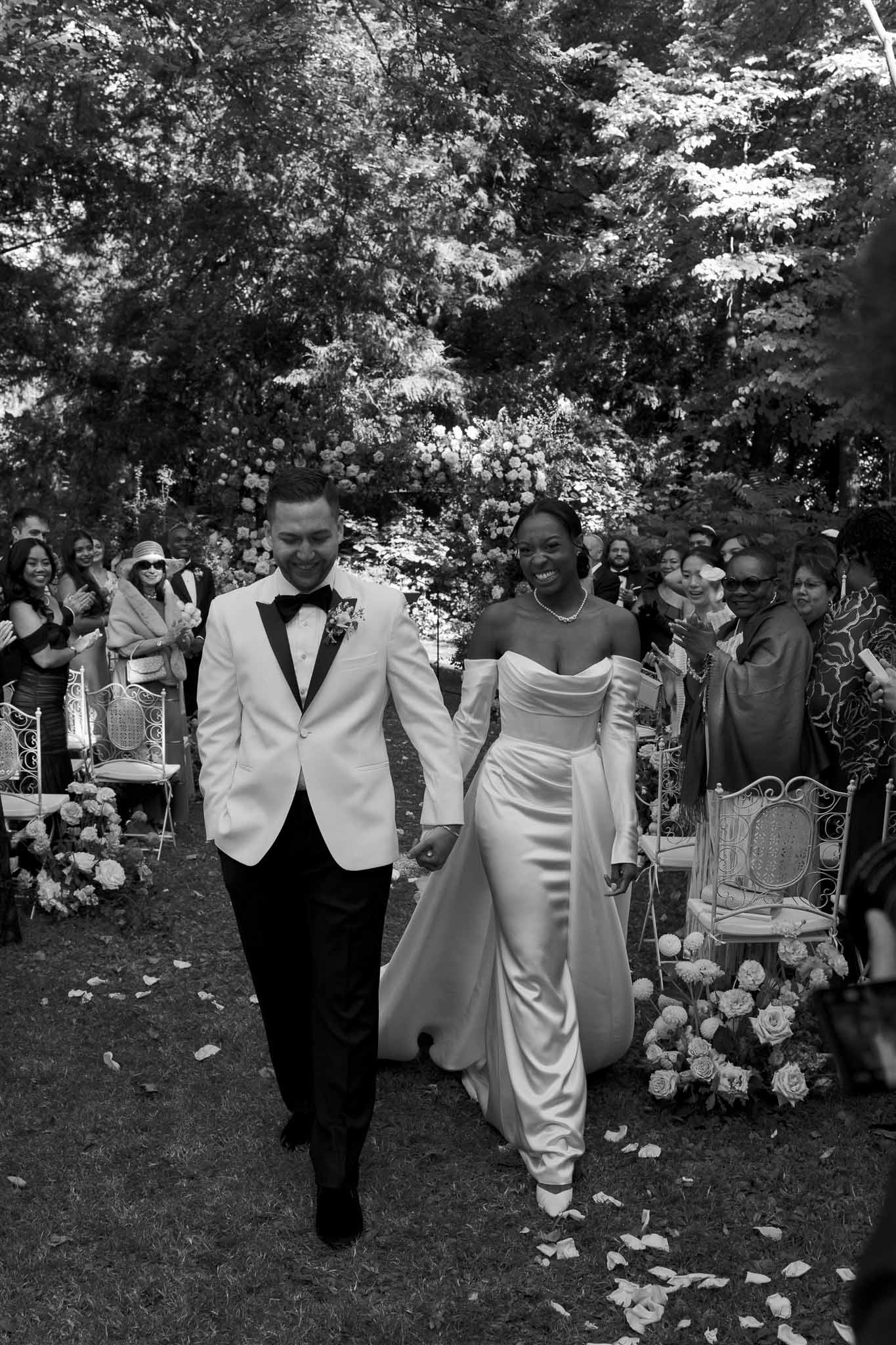 Newlywed couple walking down outdoor ceremony aisle in garden setting with guests and floral arrangements