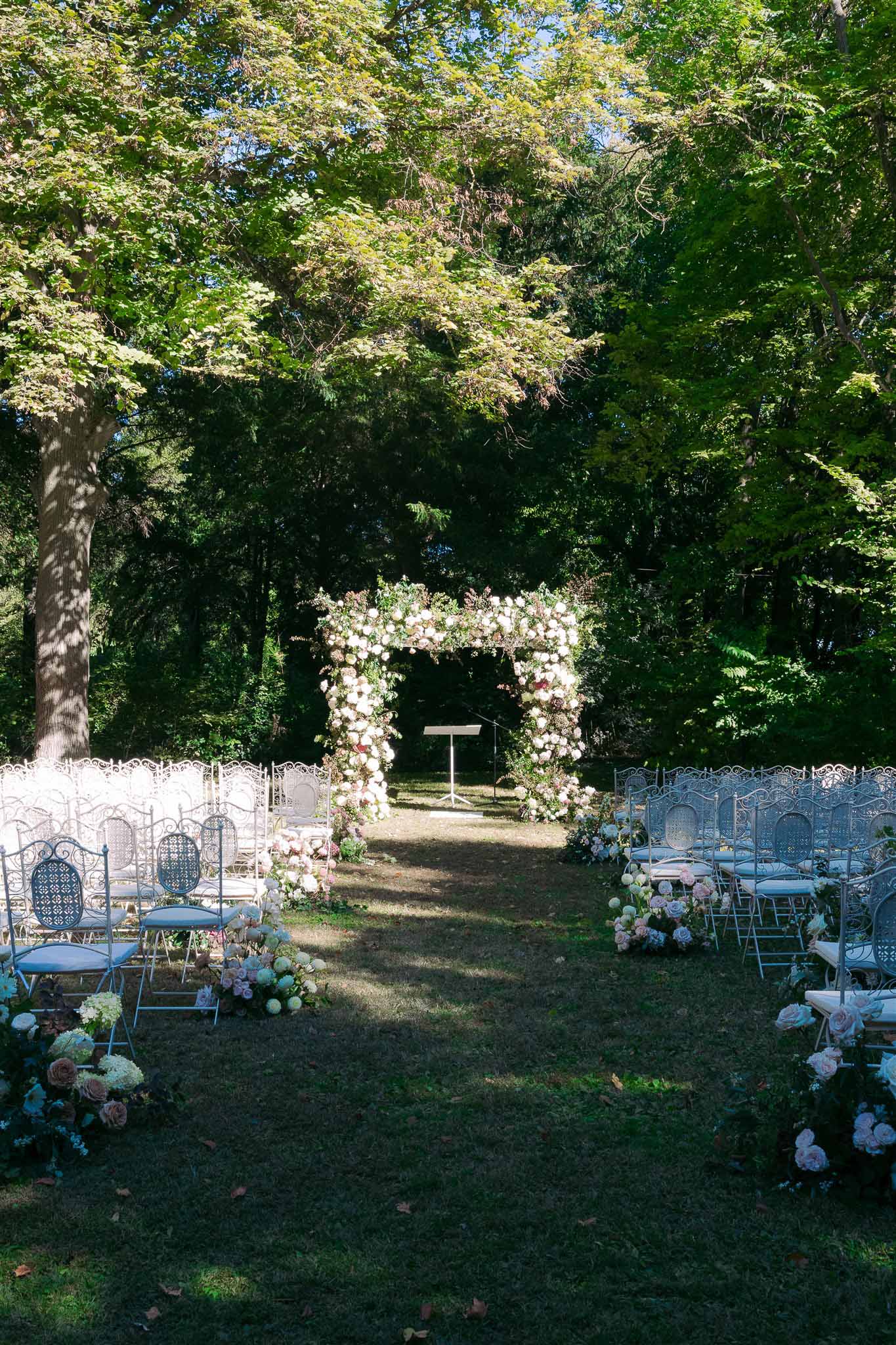 Outdoor garden ceremony setup with floral arch and wrought-iron chairs under mature trees
