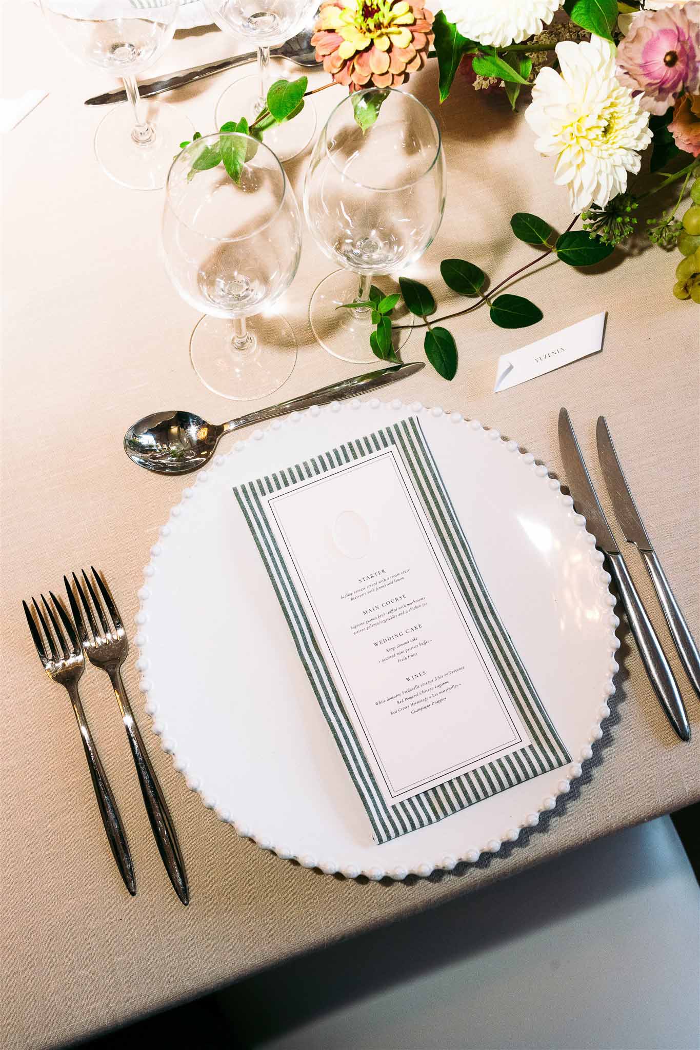 Wedding reception place setting with scalloped plate, menu card, and floral arrangement on taupe linen tablecloth