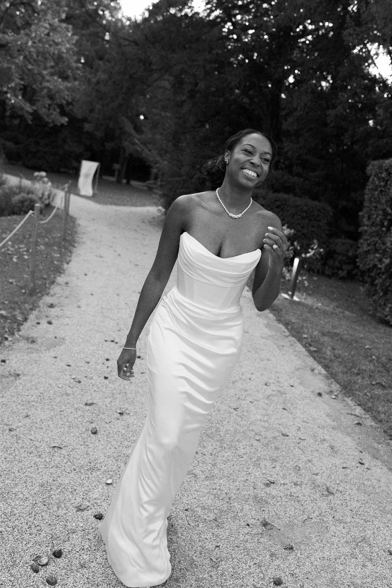 Bride in strapless wedding dress walking along tree-lined garden path in black and white portrait