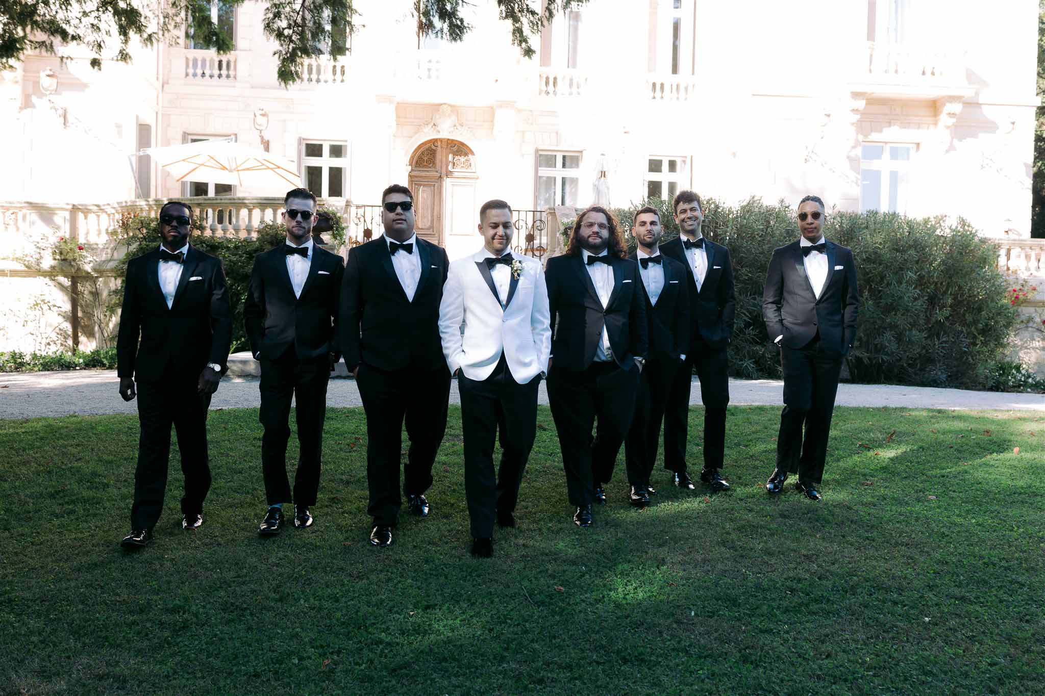 Eight groomsmen in black tuxedos and white dinner jacket groom posing at classical stone mansion courtyard