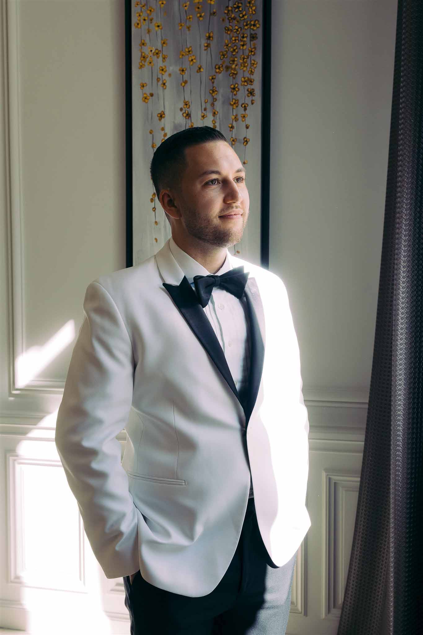 Groom in white dinner jacket and bow tie posing by window during wedding preparation