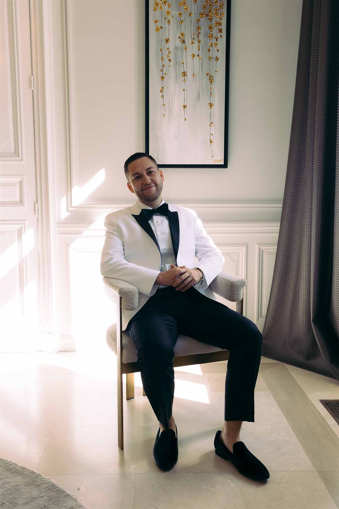 Groom in cream jacket and bow tie seated in elegant indoor setting during wedding preparations