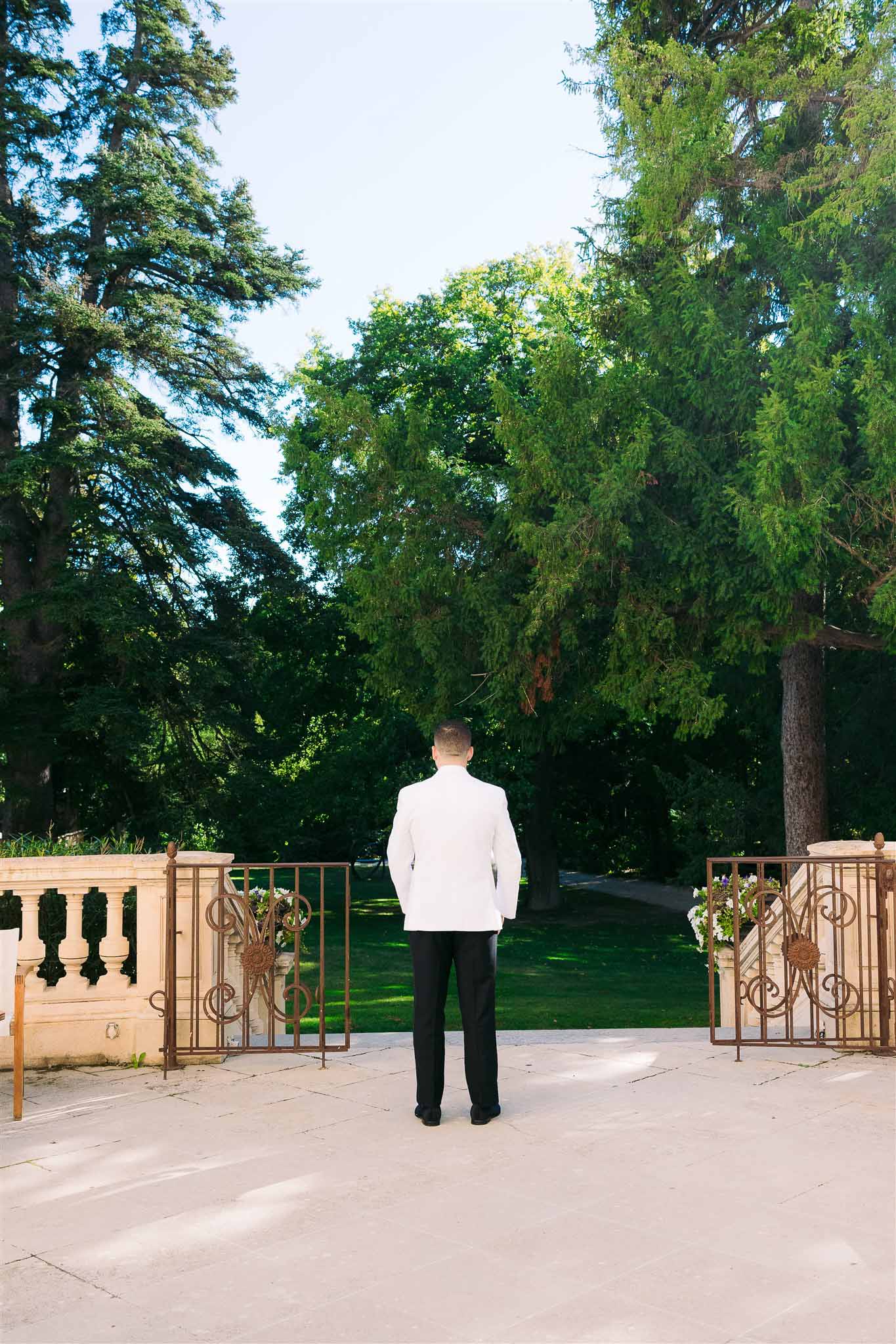 Groom in white jacket standing before ornamental gates at formal garden venue