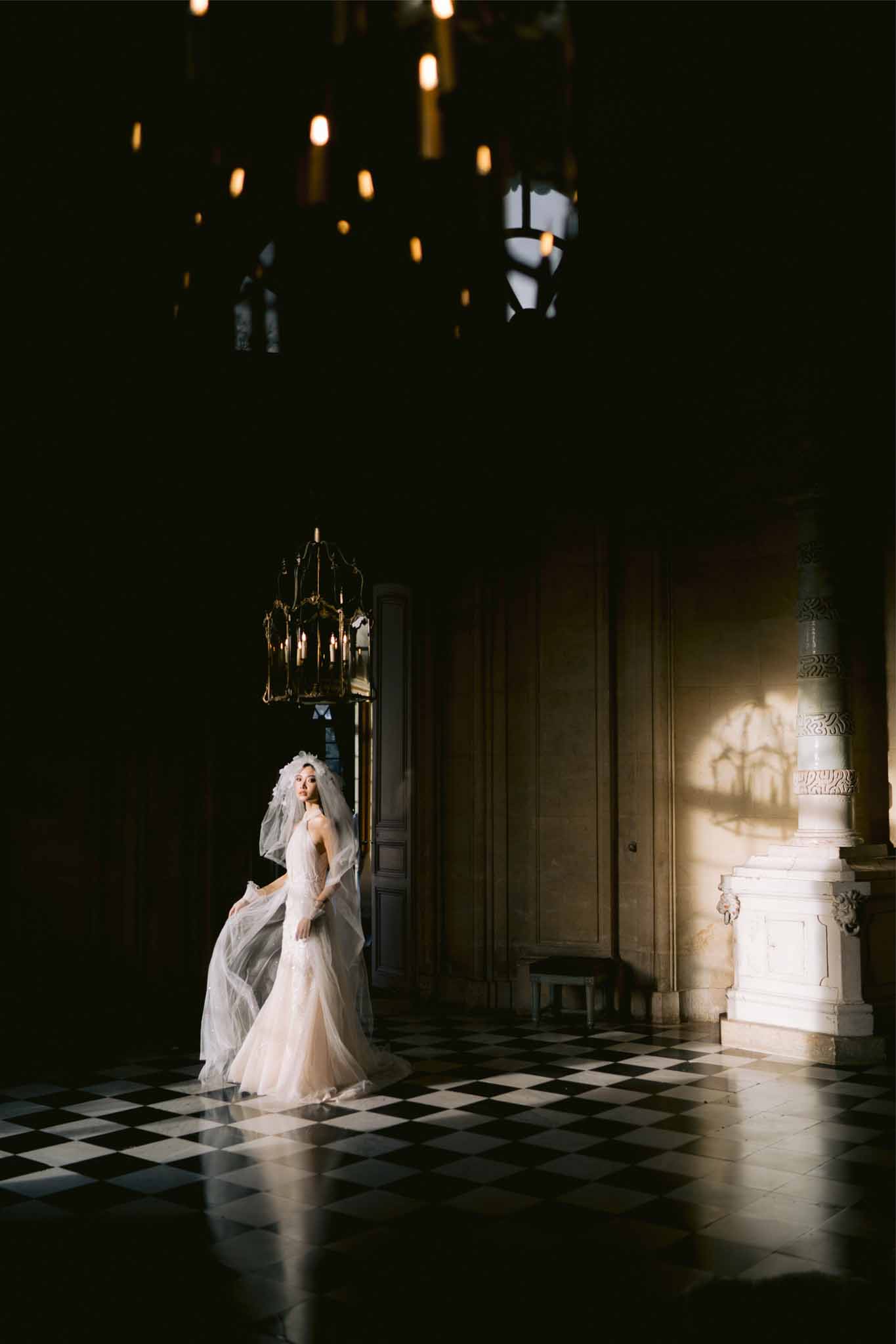 Bride in ivory gown with veil standing in classical interior hallway with checkered floor and lantern lighting
