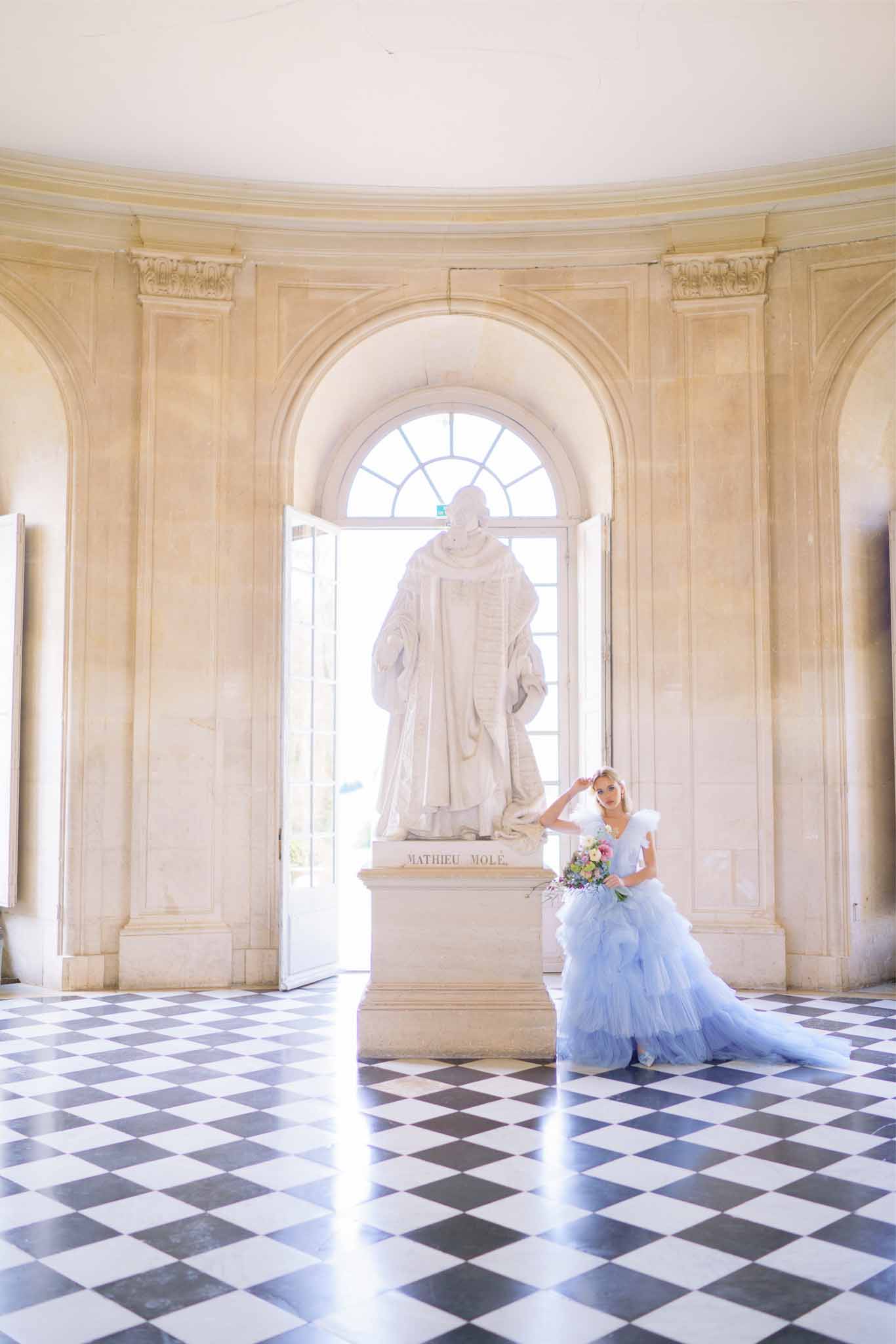 Bride in blue tulle gown with bouquet poses beside marble statue in classical palace interior