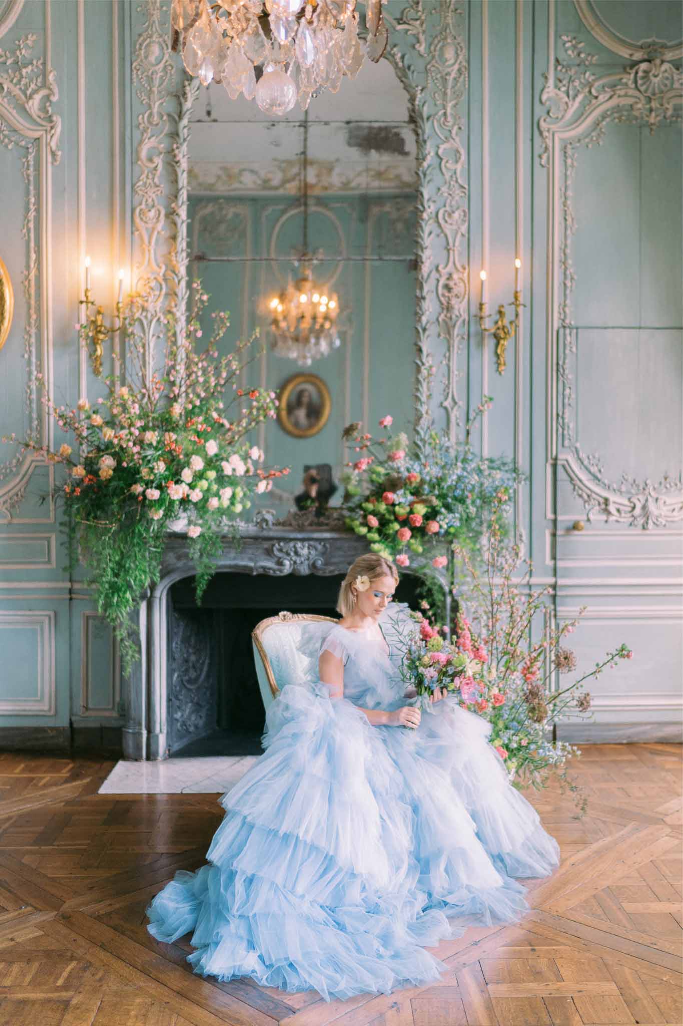 Bride in blue ballgown with bouquet in ornate neoclassical ballroom with marble fireplace