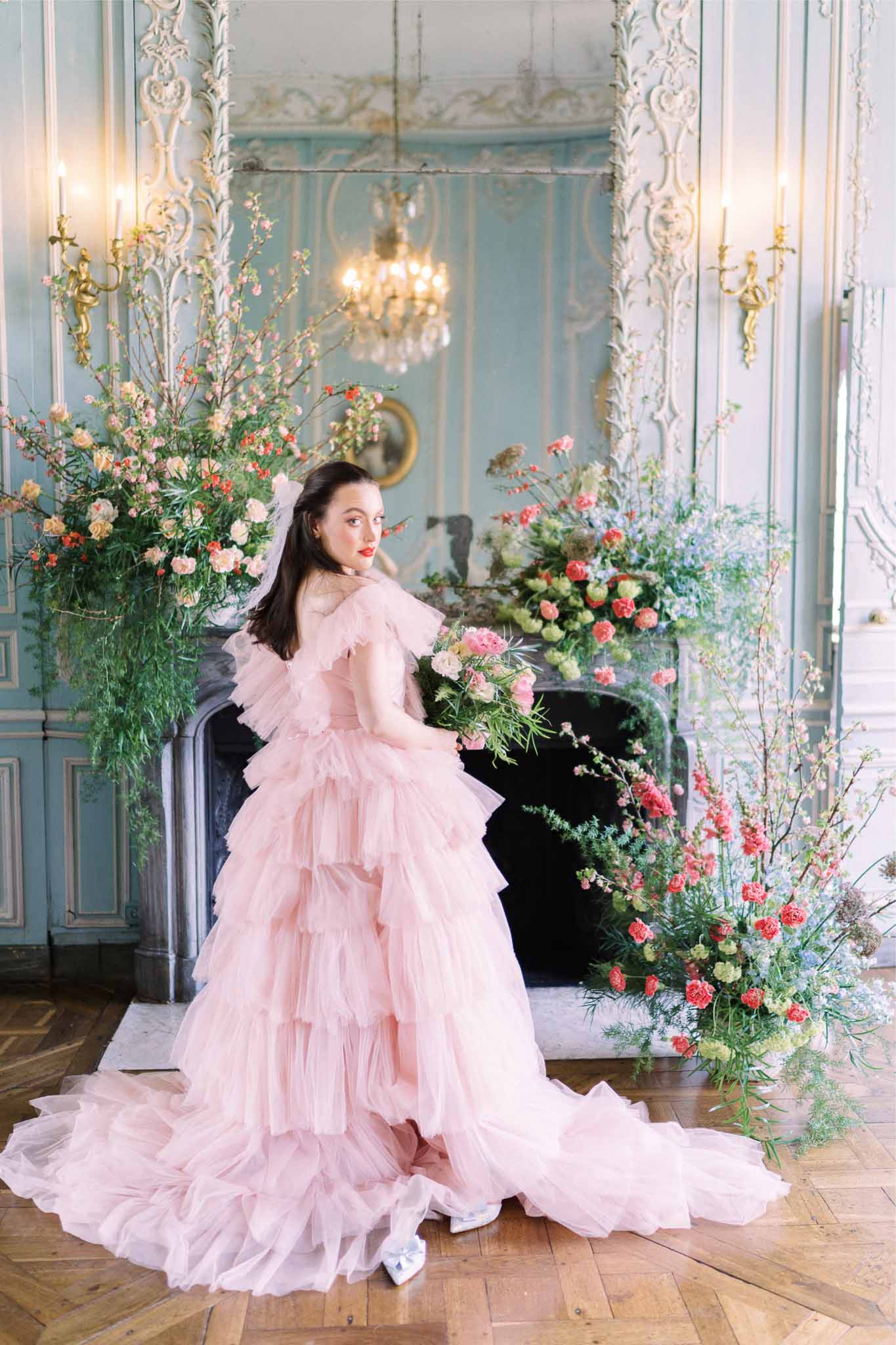 Bride in pale pink gown posing in ornate neoclassical ballroom with crystal chandelier and floral arrangements