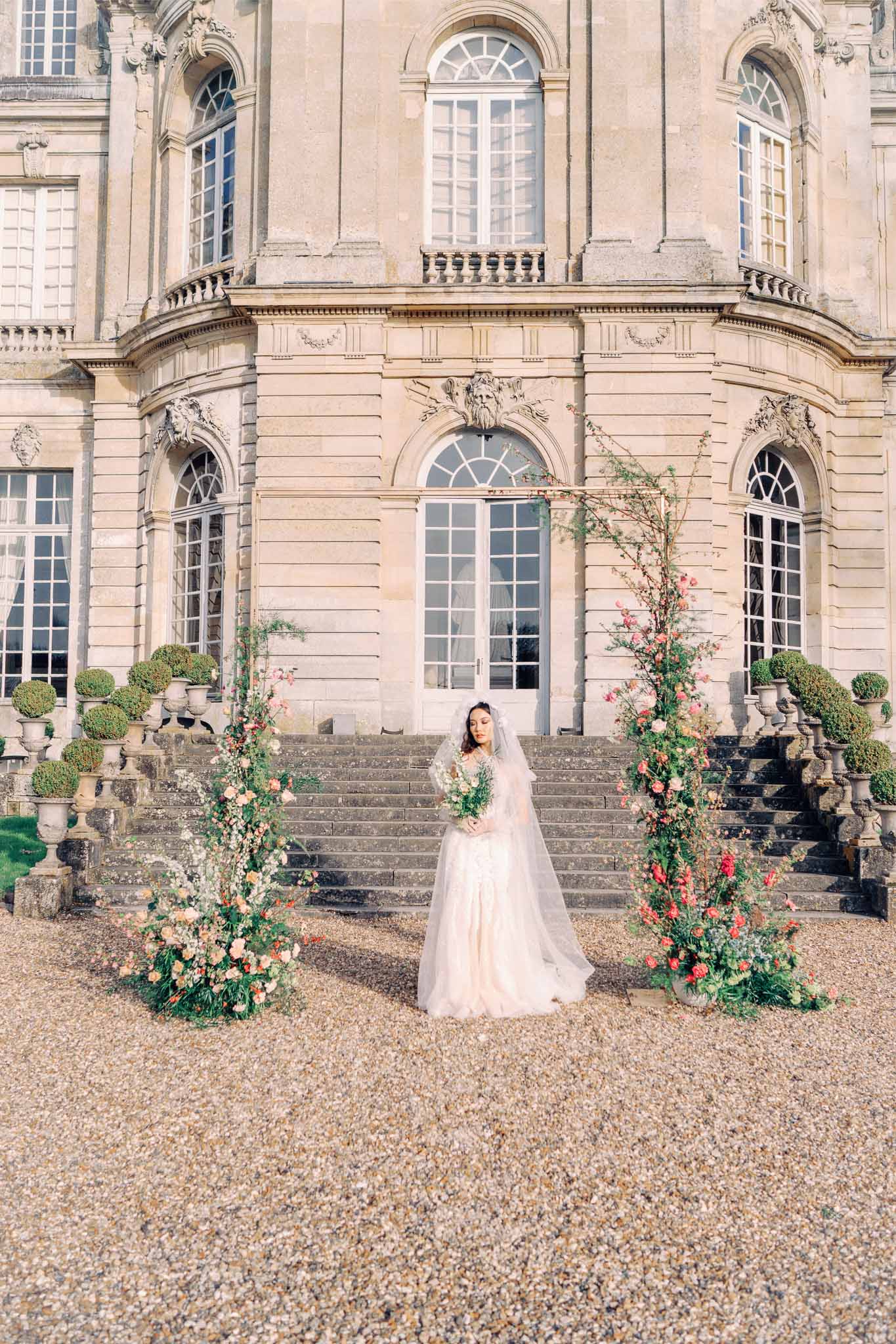 Bride in ivory gown with bouquet standing before classical stone mansion with floral installations