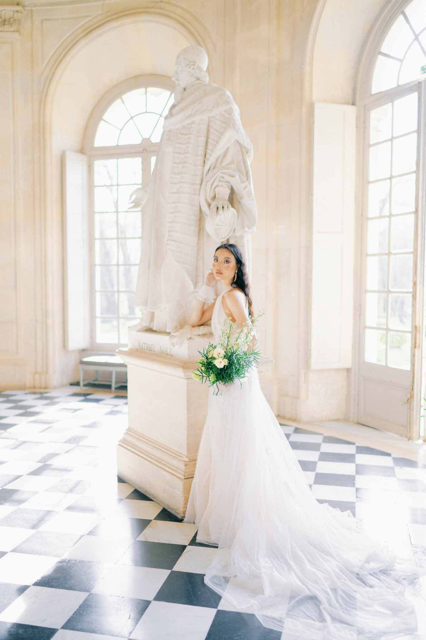 Bride in ivory gown with bouquet standing in neoclassical gallery with arched windows and marble statue