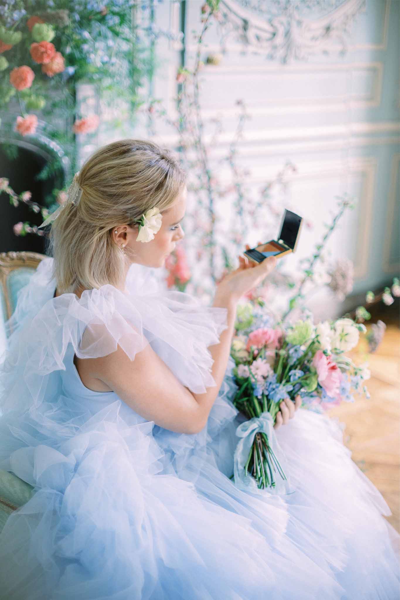 Bride in pale blue gown holding mirror compact during getting ready preparations indoors