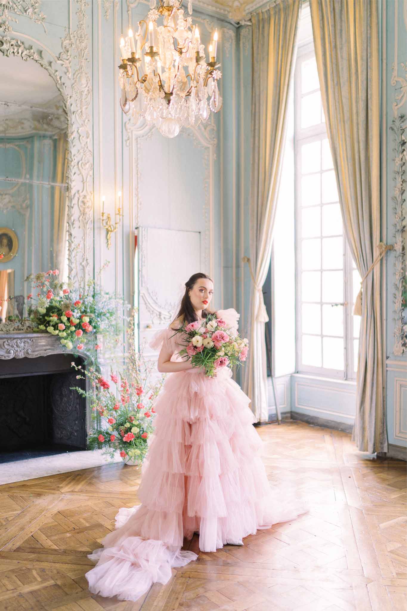 Bride in blush pink gown holding coral rose bouquet in ornate palace ballroom with crystal chandelier