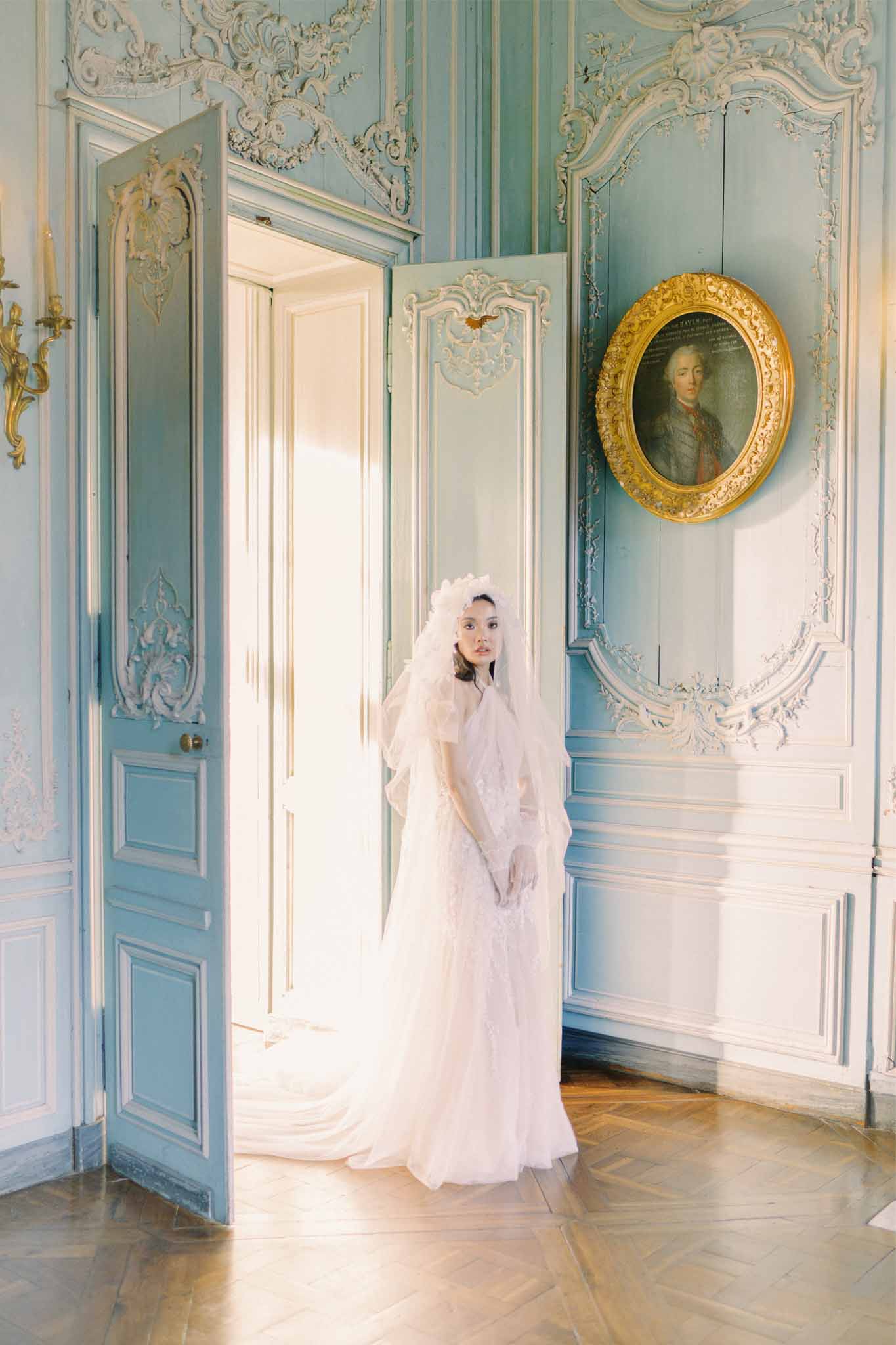 Bride in ivory gown standing in ornate neoclassical room with rococo paneling and historical portraits