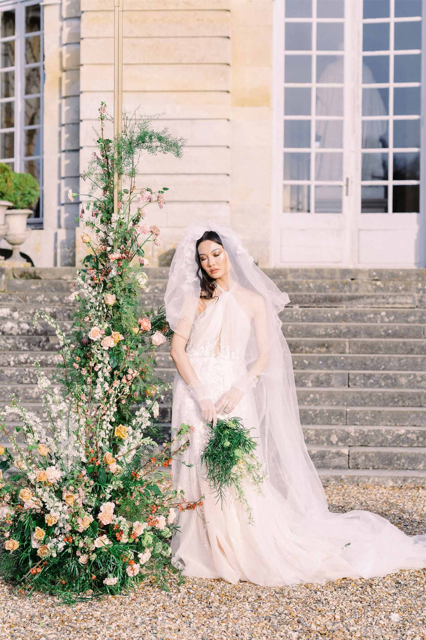 Bride in ivory gown with peach bouquet poses at classical stone building courtyard